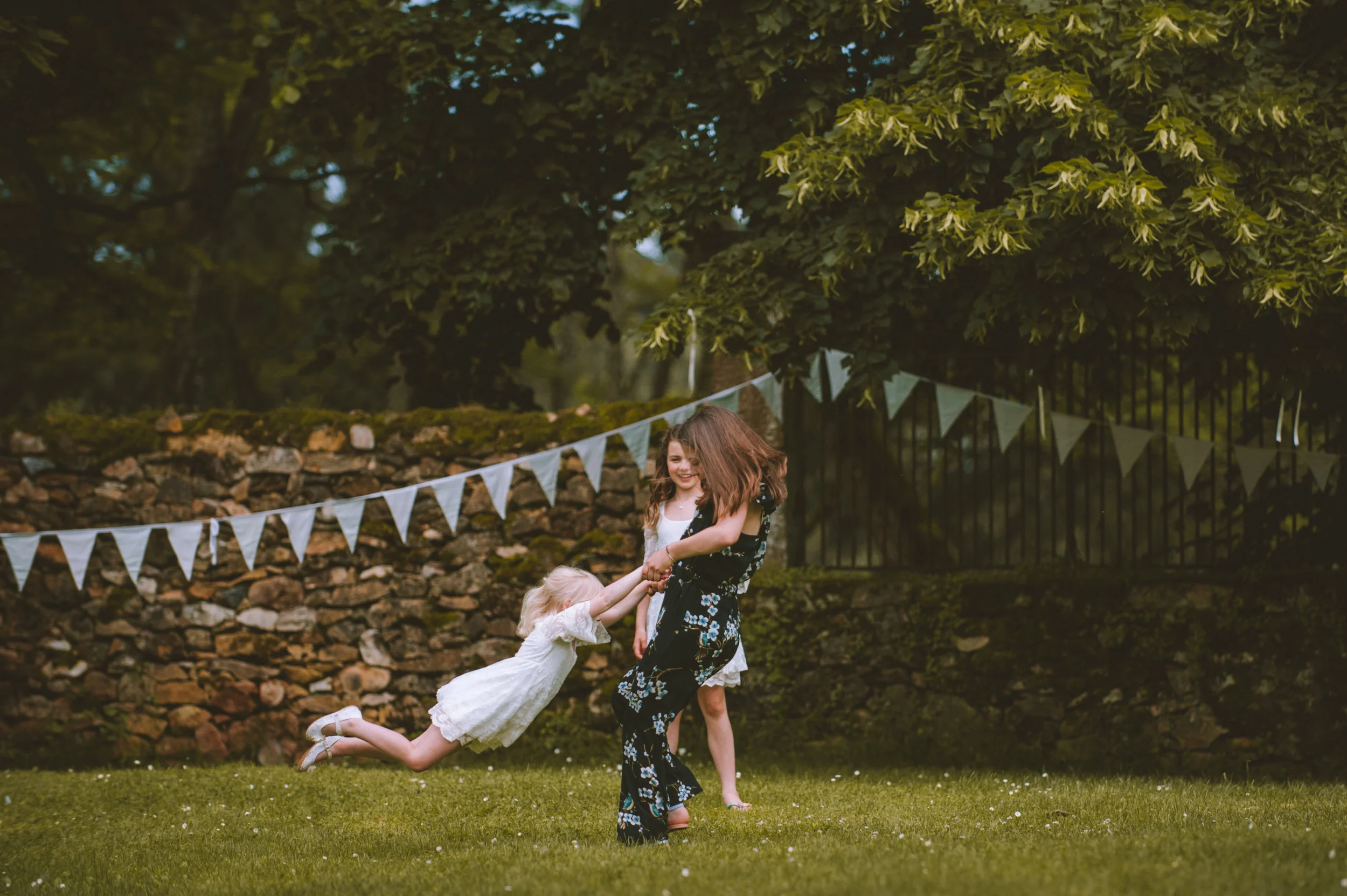 Three young girls playing tug of war with a stick outdoors, with greenery and bunting in the background.