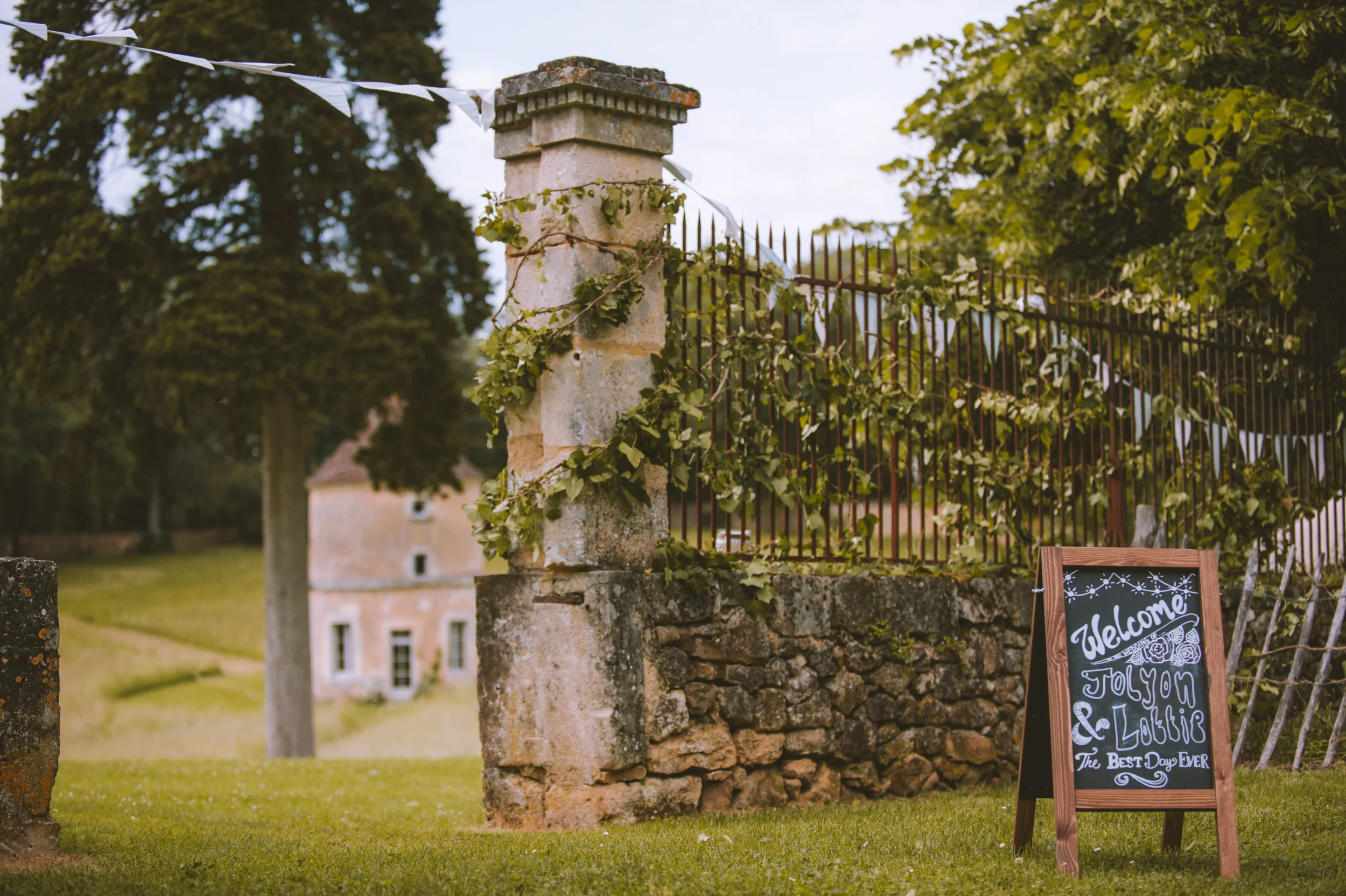 A decorative signboard welcoming guests, situated on a lawn outside a stone and iron fence, leading toward a historic building in the background.