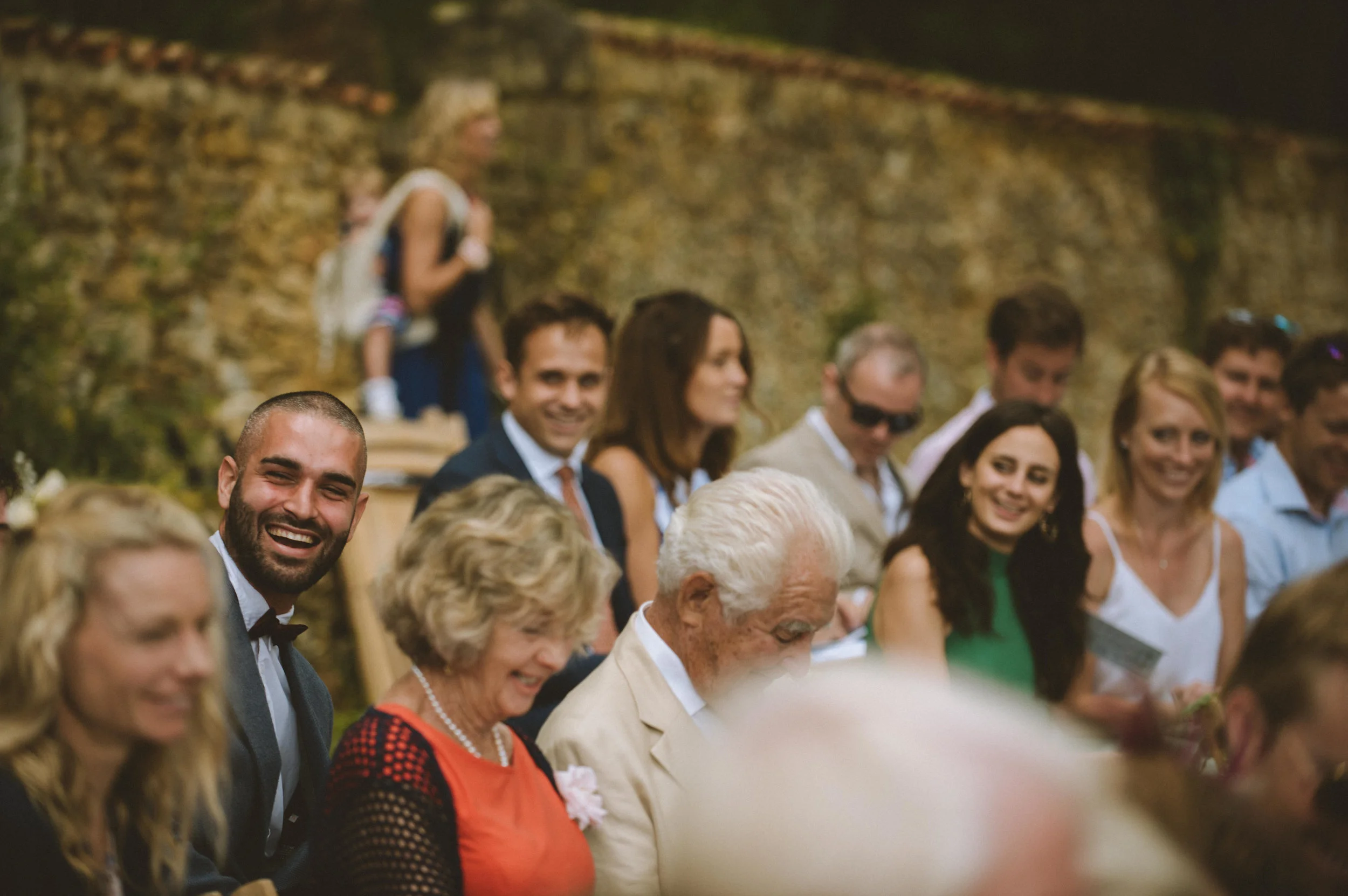 Group of people attending a wedding or celebration, seated and smiling in front of a stone wall.