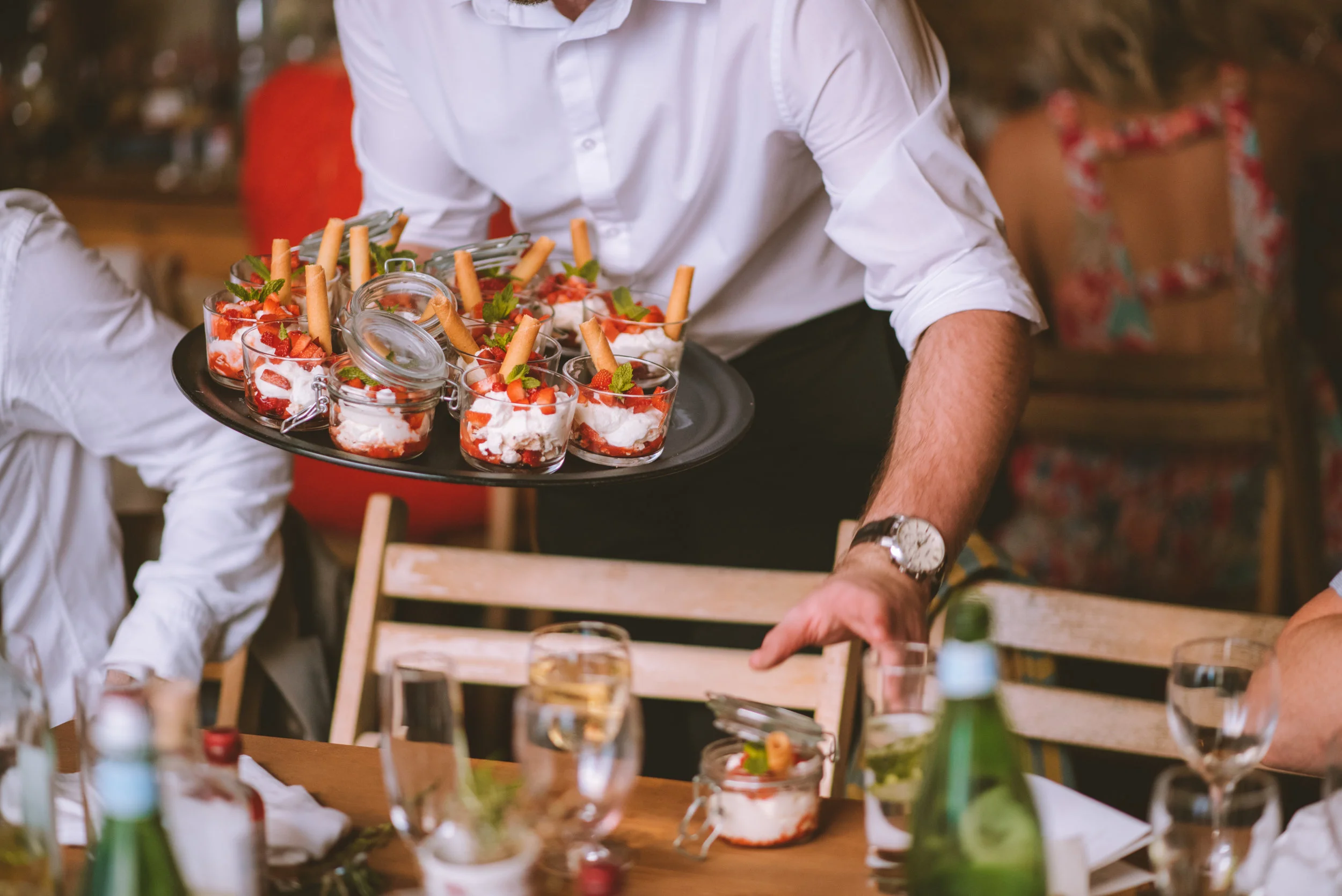 Person serving dessert cups with strawberries and whipped cream at a table with glasses and bottles in the background.