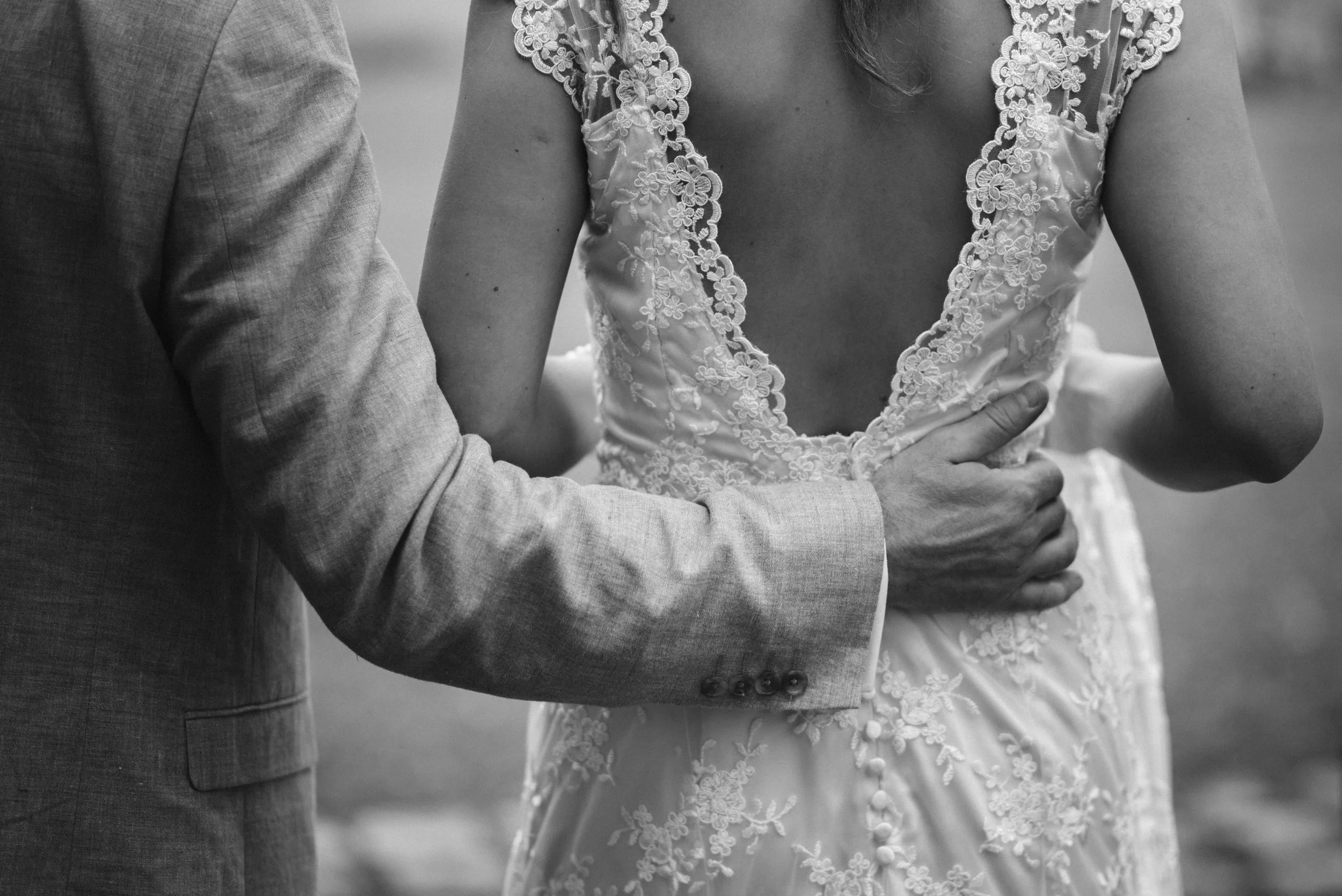 Close-up of a groom holding a bride by the waist during their wedding, focusing on their arms and the back of her lace wedding dress.