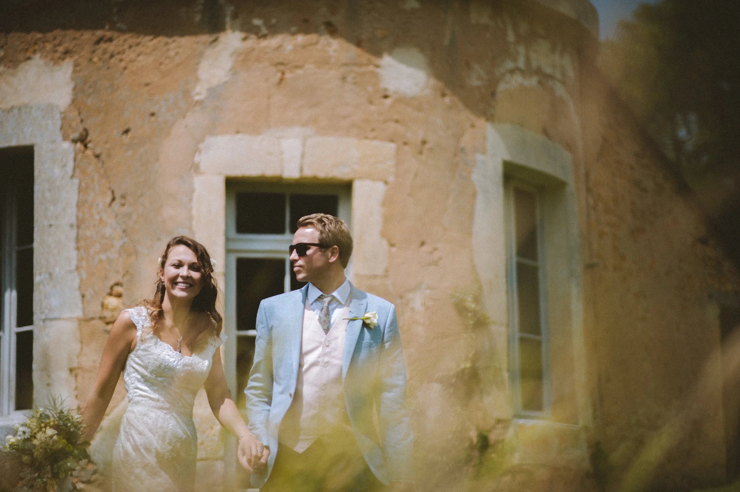 A bride and groom walking hand in hand outdoors in front of an old stone building during a wedding.