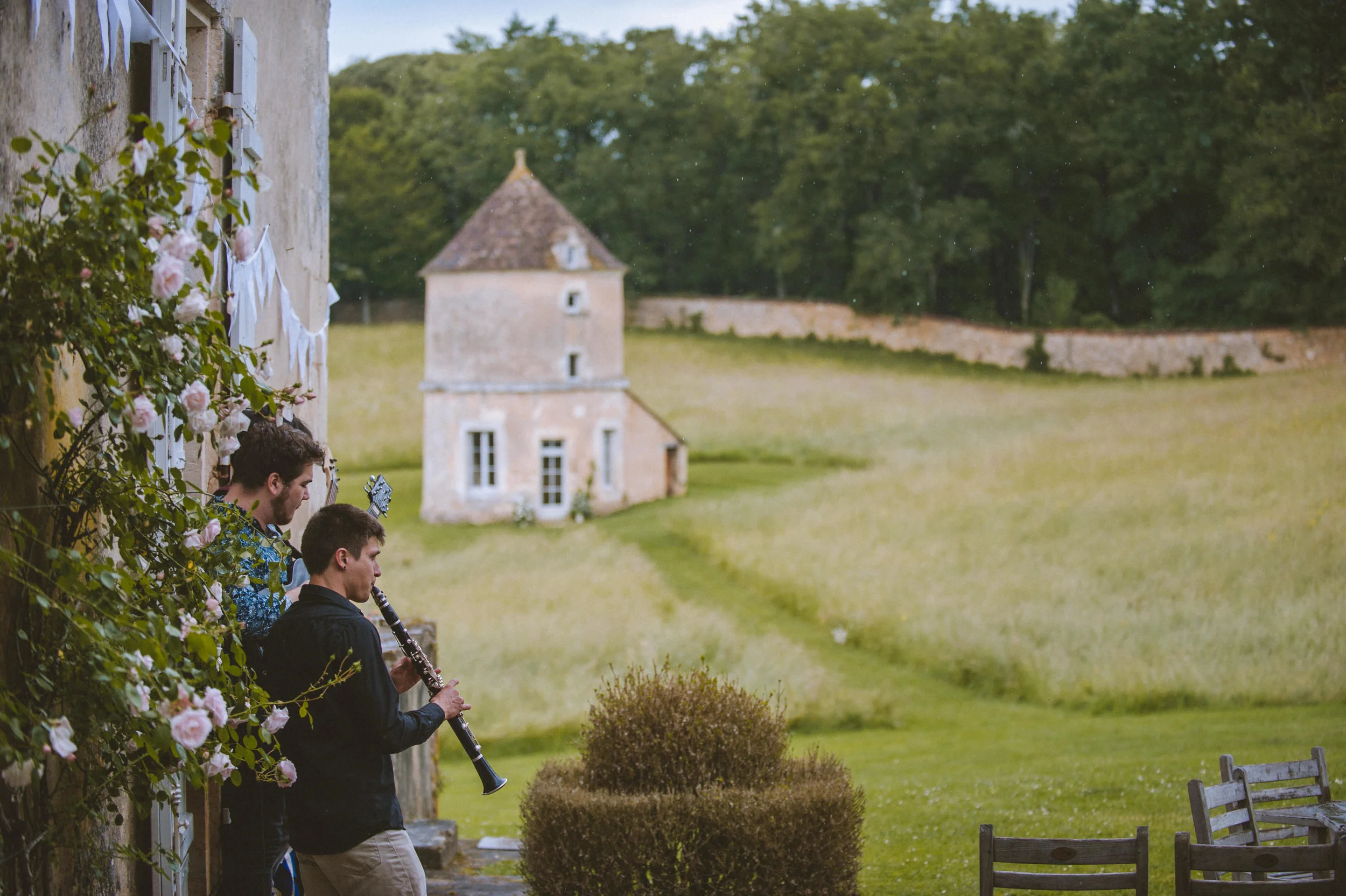 Two young men stand on a porch next to a flowering bush, one playing a clarinet while the other watches, with a scenic green field and a small tower in the background.