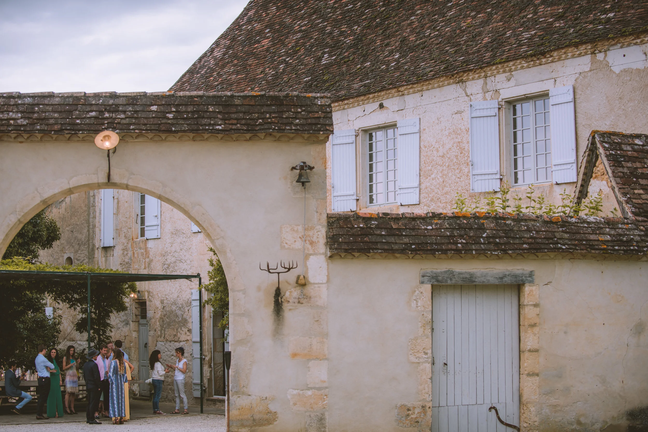 Group of people gathered and chatting outside in a courtyard surrounded by old stone buildings with white shutters and tiled roofs.