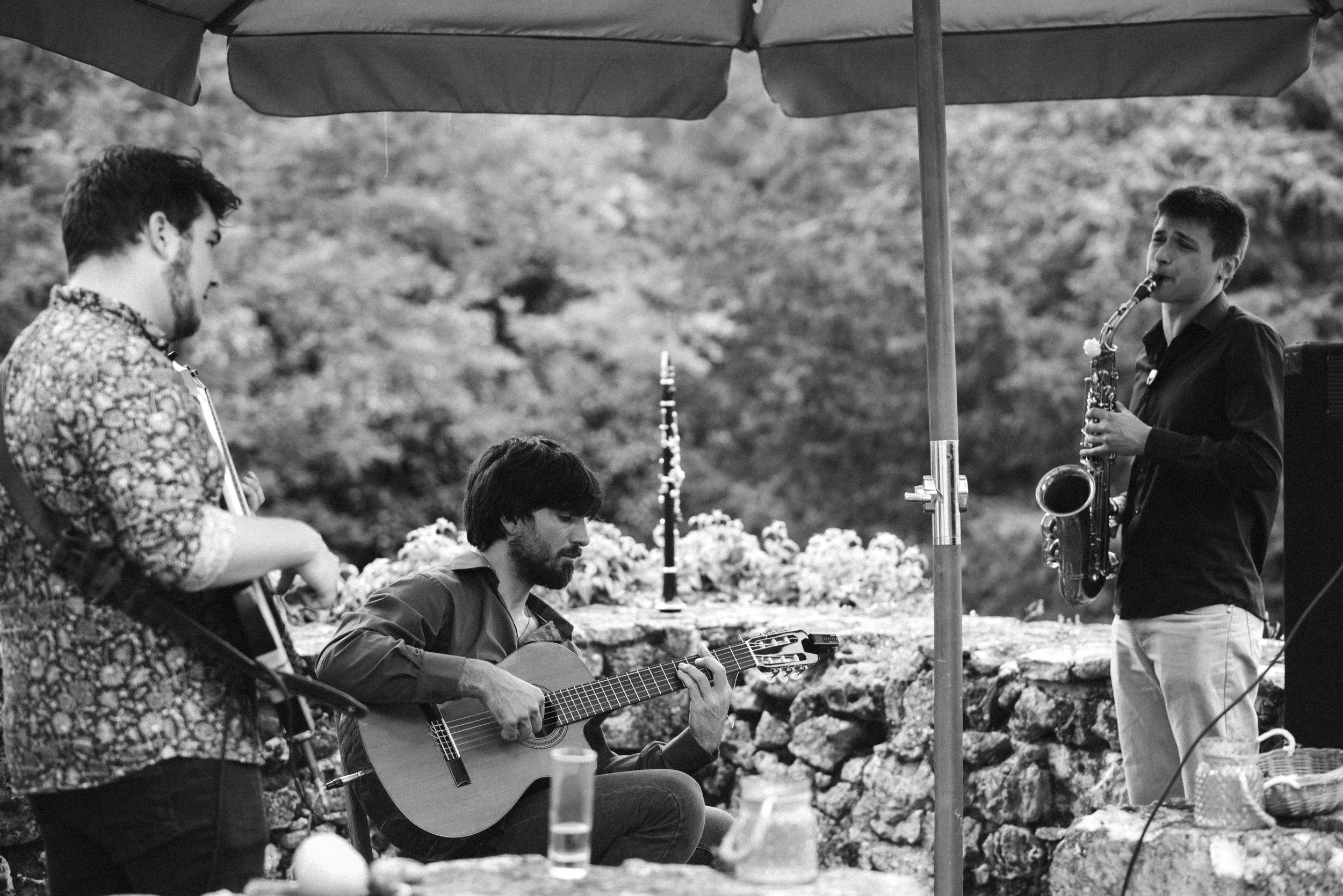 Three musicians performing outdoors under a large umbrella. One playing an acoustic guitar, another playing a saxophone, and the third possibly playing a percussion instrument. The background shows trees and a stone wall.