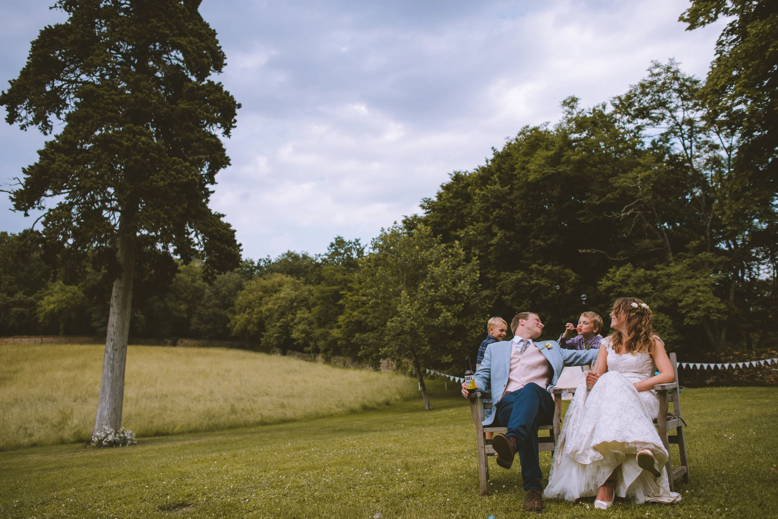 A newlywed couple sitting on a bench outdoors with two children, in a spacious green field with trees, during daytime with cloudy sky.