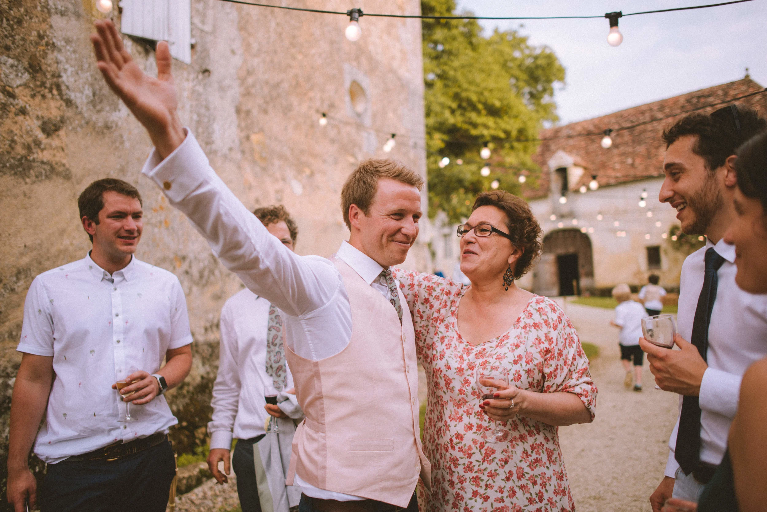 Group of people celebrating outdoors during a social gathering, with a man raising his hand and a woman holding a drink, in a rustic outdoor setting with string lights and a stone wall.