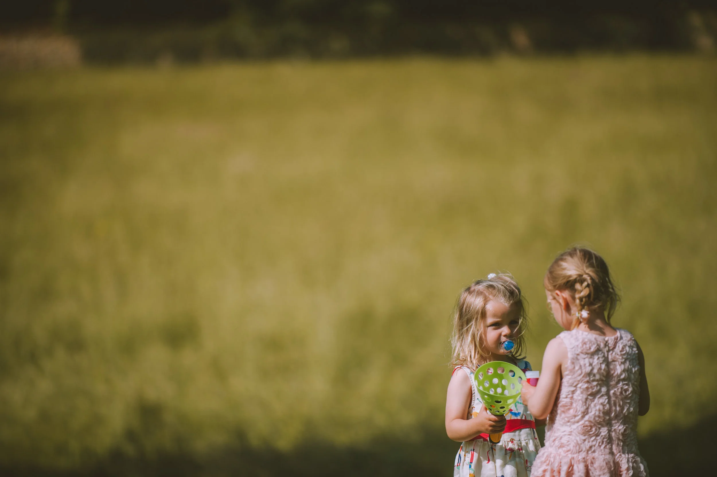 Two young girls playing outdoors near a body of water, one with a pacifier, holding a green scoop, talking to the other girl in a pink dress.