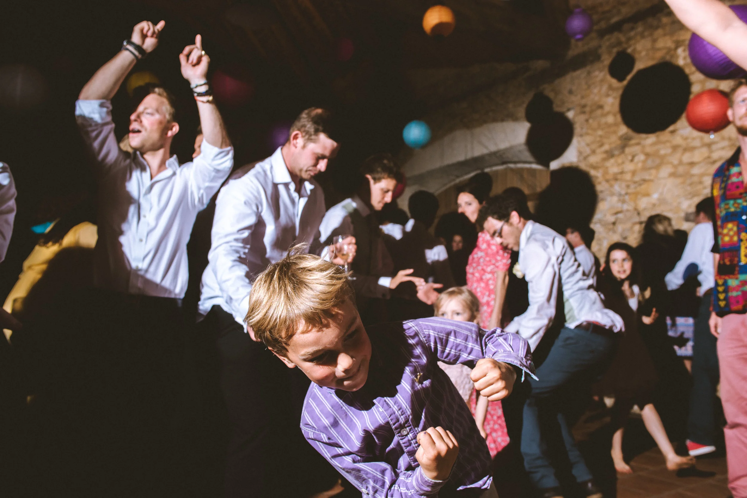 People dancing at a party or celebration in a rustic venue with stone walls and colorful lanterns overhead.