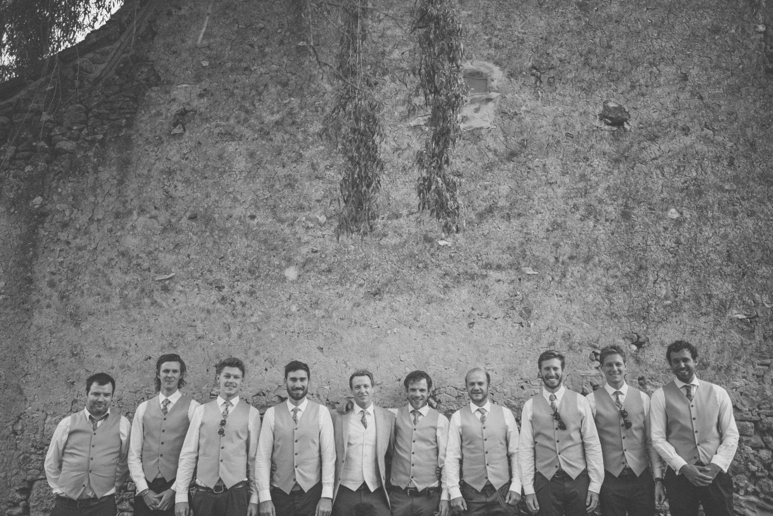 A group of ten men dressed in vests and dress shirts standing outdoors, with an old stone wall in the background.