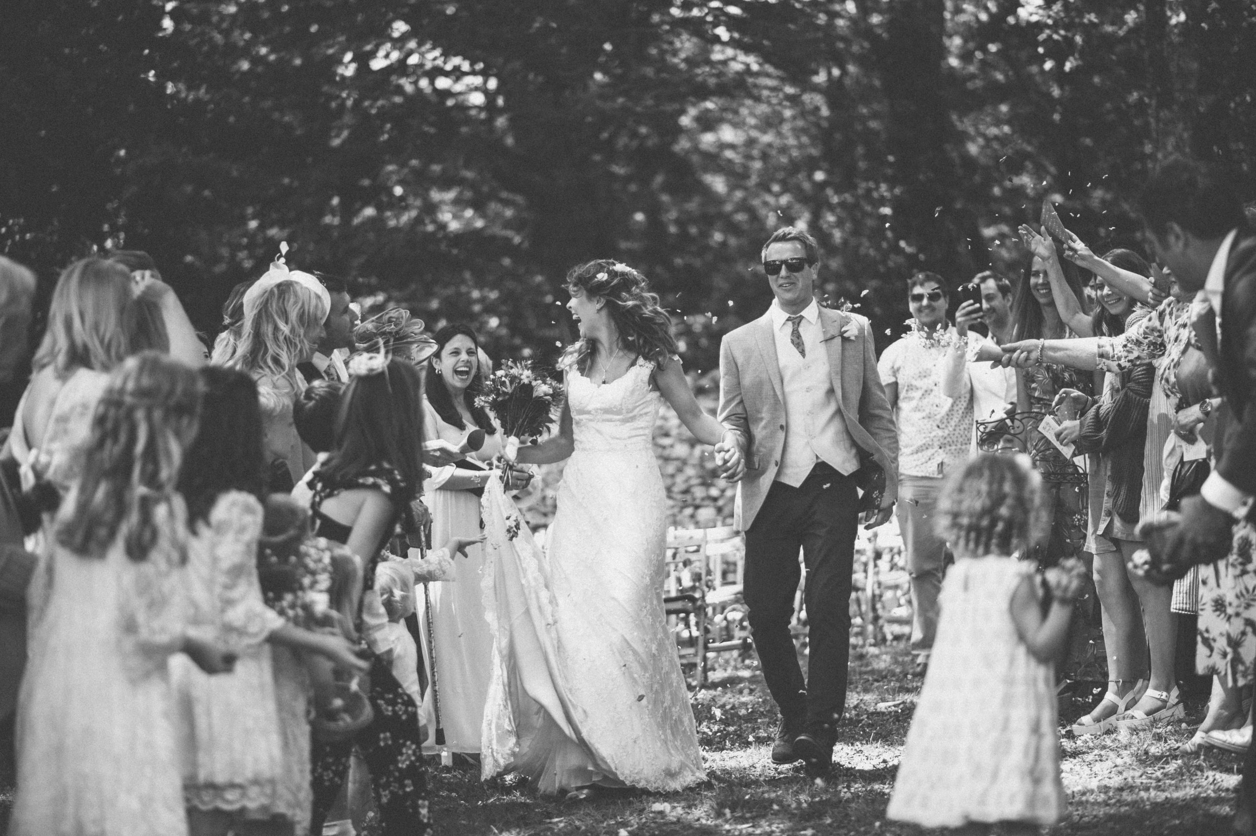 Black and white photo of a bride and groom walking hand-in-hand through a crowd of smiling guests outdoors. The bride wears a wedding gown, and the groom is dressed in a suit with sunglasses. Guests on both sides cheer and throw confetti.