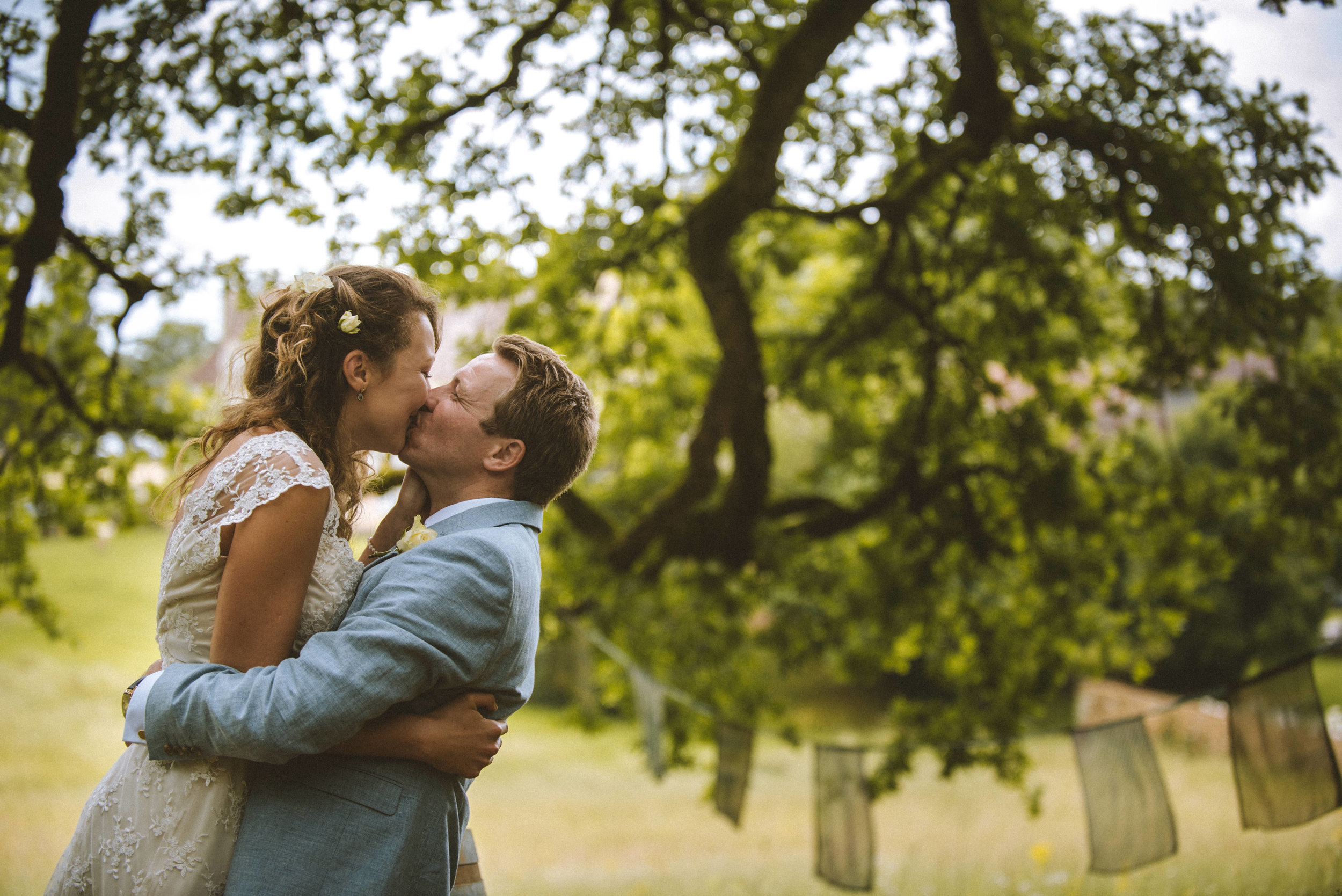 A bride and groom sharing a kiss outdoors during a wedding celebration, with trees and fabric decorations in the background.
