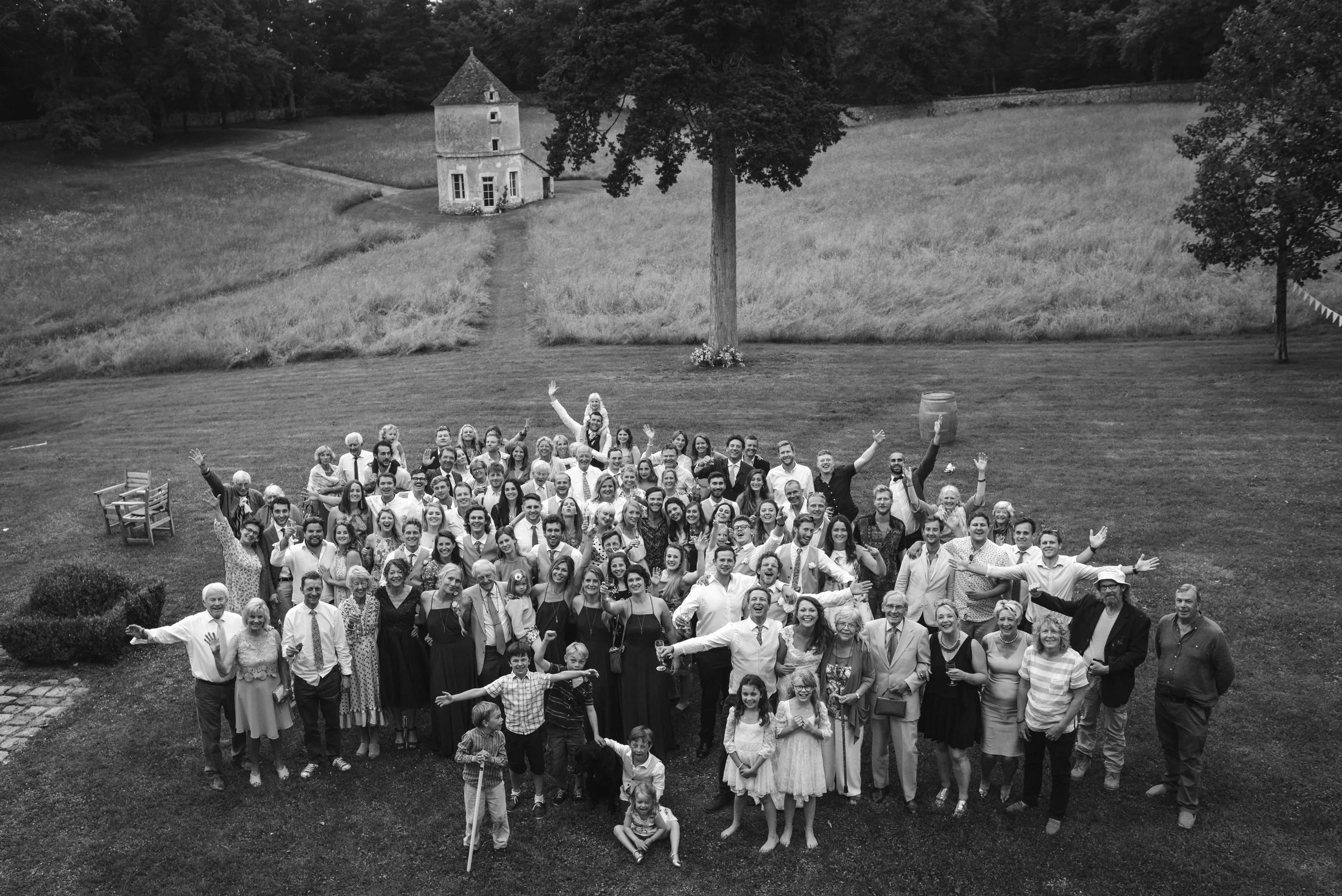 A large group of people gathered outdoors on a grassy field, posing for a group photo. The setting includes a large tree, a barrel, benches, and a small tower in the background, with a rural landscape.