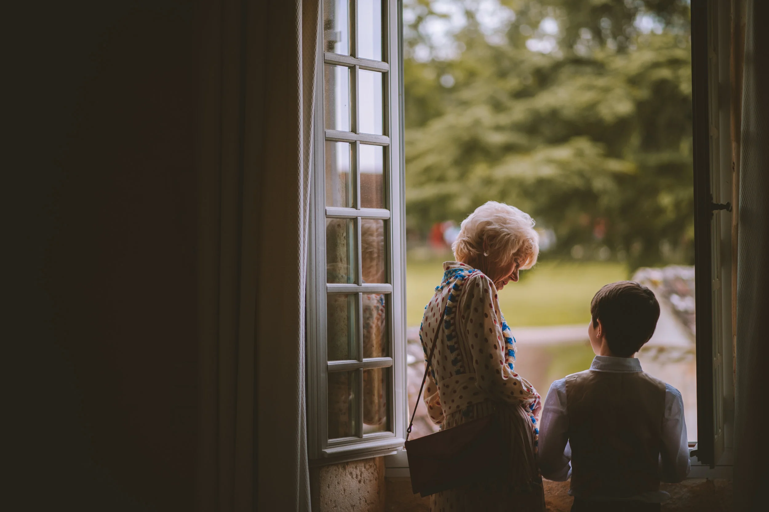 An elderly woman and a young boy stand at an open window, looking outside at a green, tree-filled area with parked cars in the background.
