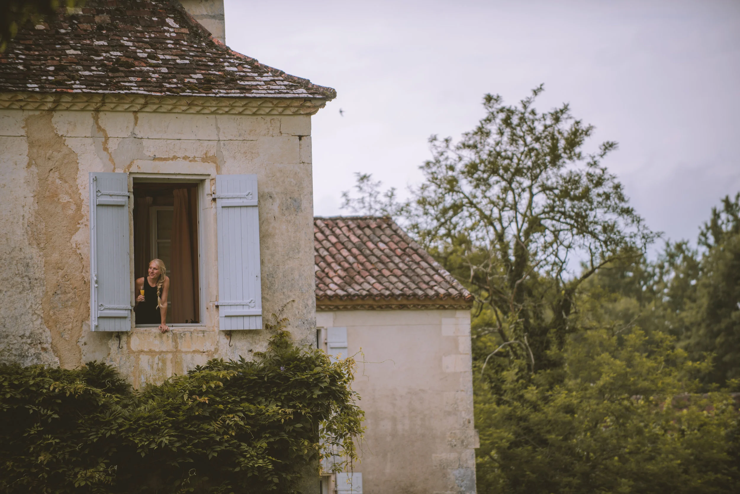 A woman leaning out of a second-story window of an old stone building, holding a glass of wine, with green foliage and trees in the background.