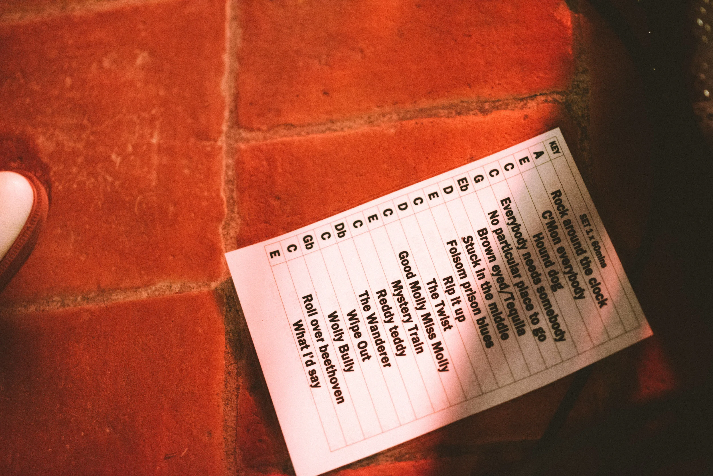 A printed song setlist on a piece of paper resting on a red brick floor.