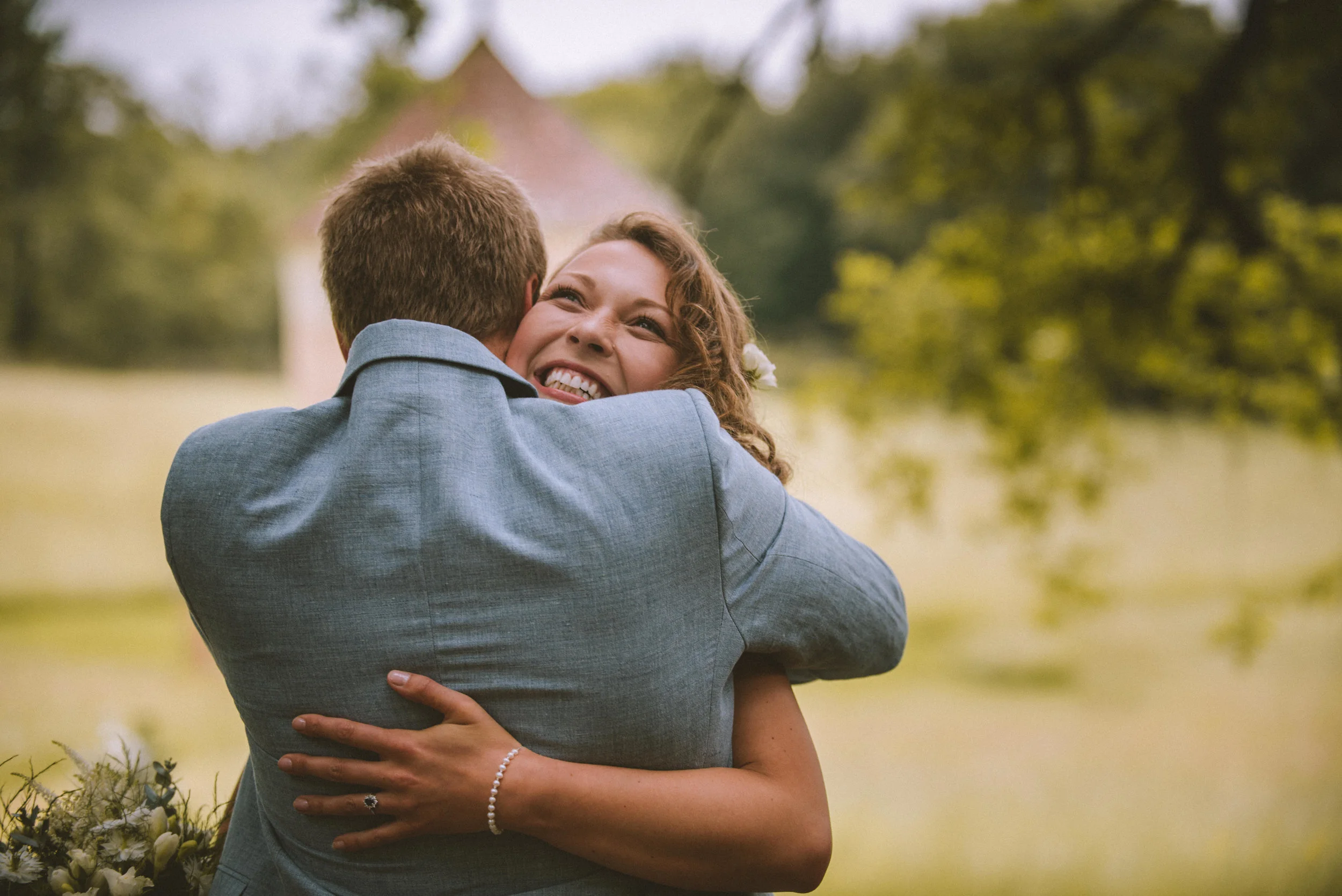 A woman with curly hair and smiling face hugging a man in a coat, outdoors in a green, tree-filled area.
