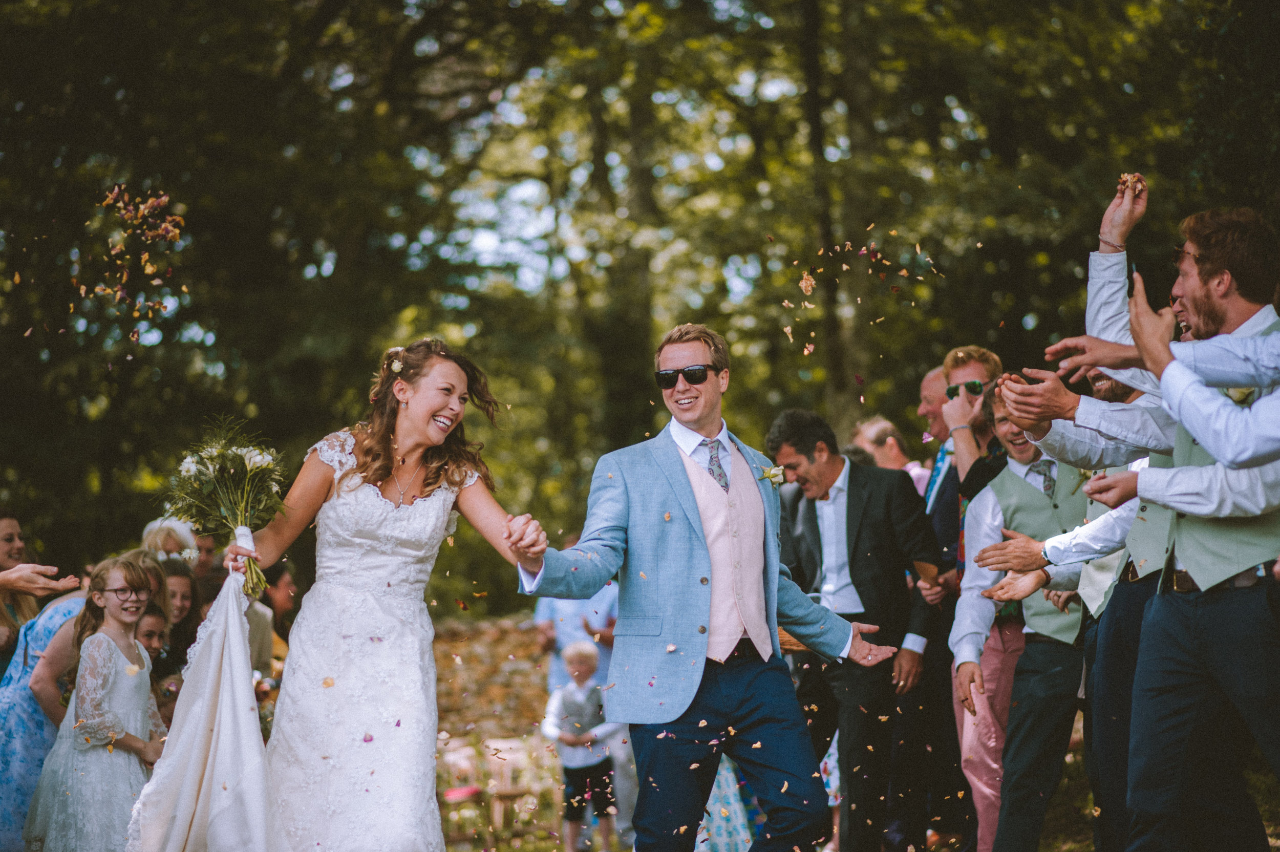 A bride and groom celebrating outdoors with guests, confetti in the air, trees in the background, and everyone smiling.