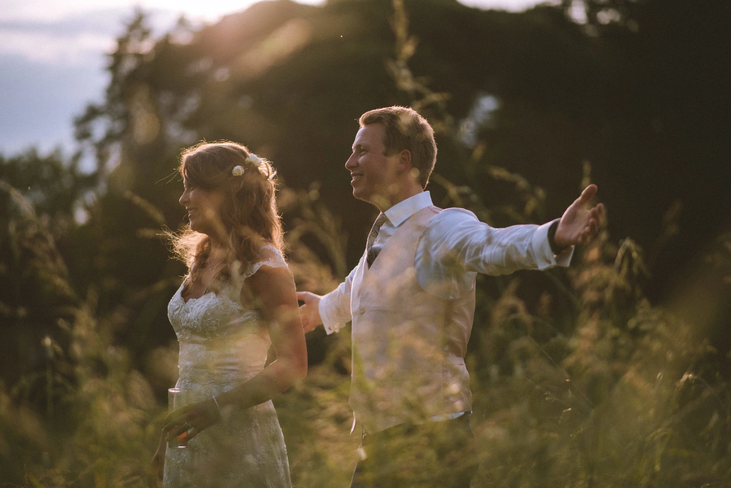 A man and woman, dressed in wedding attire, enjoying a moment outdoors with the man spreading his arms wide and the woman smiling, during sunset in a natural setting.