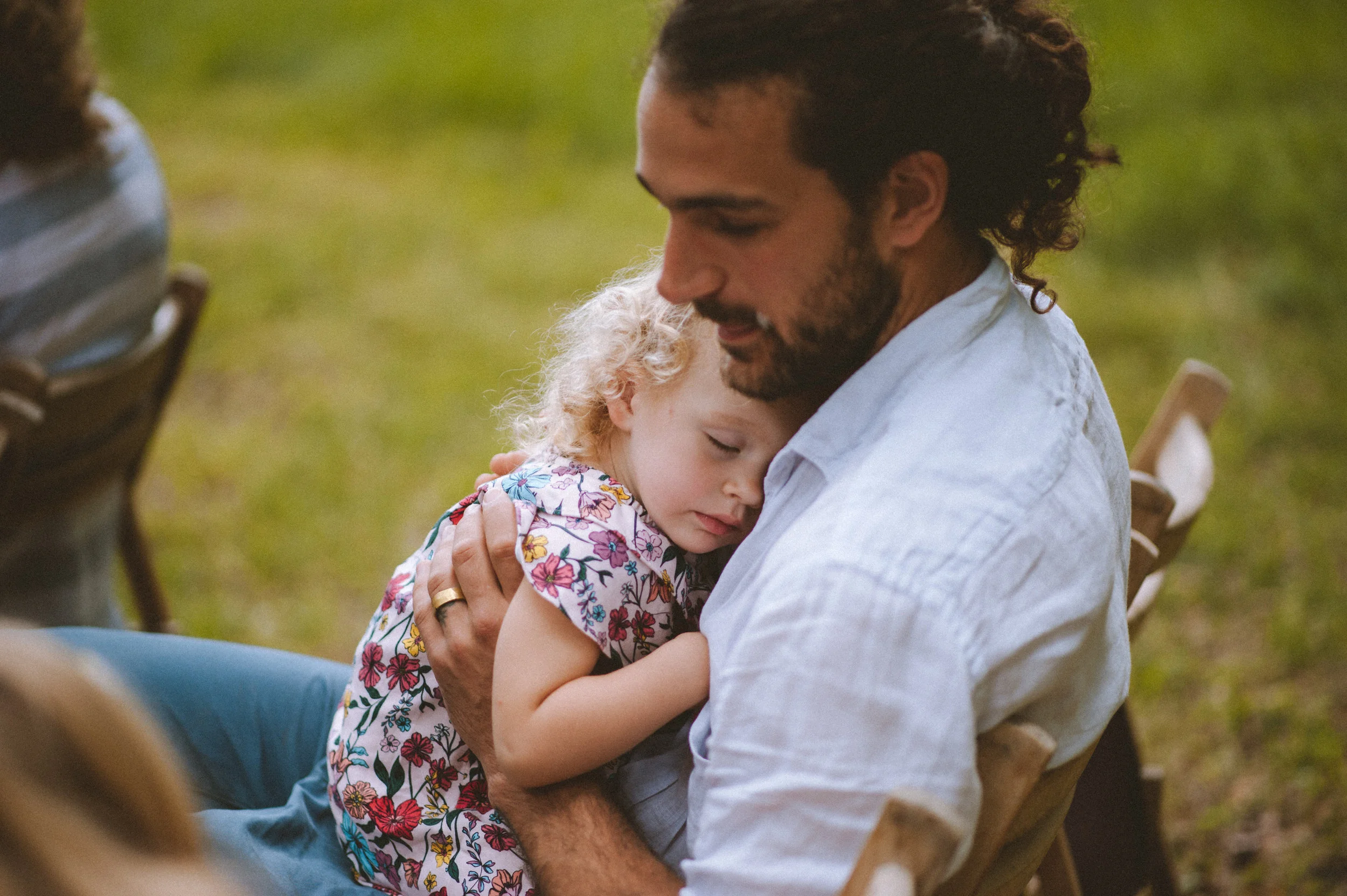 A man holding a sleeping young girl with curly blonde hair and a floral dress, sitting outdoors on a wooden chair.