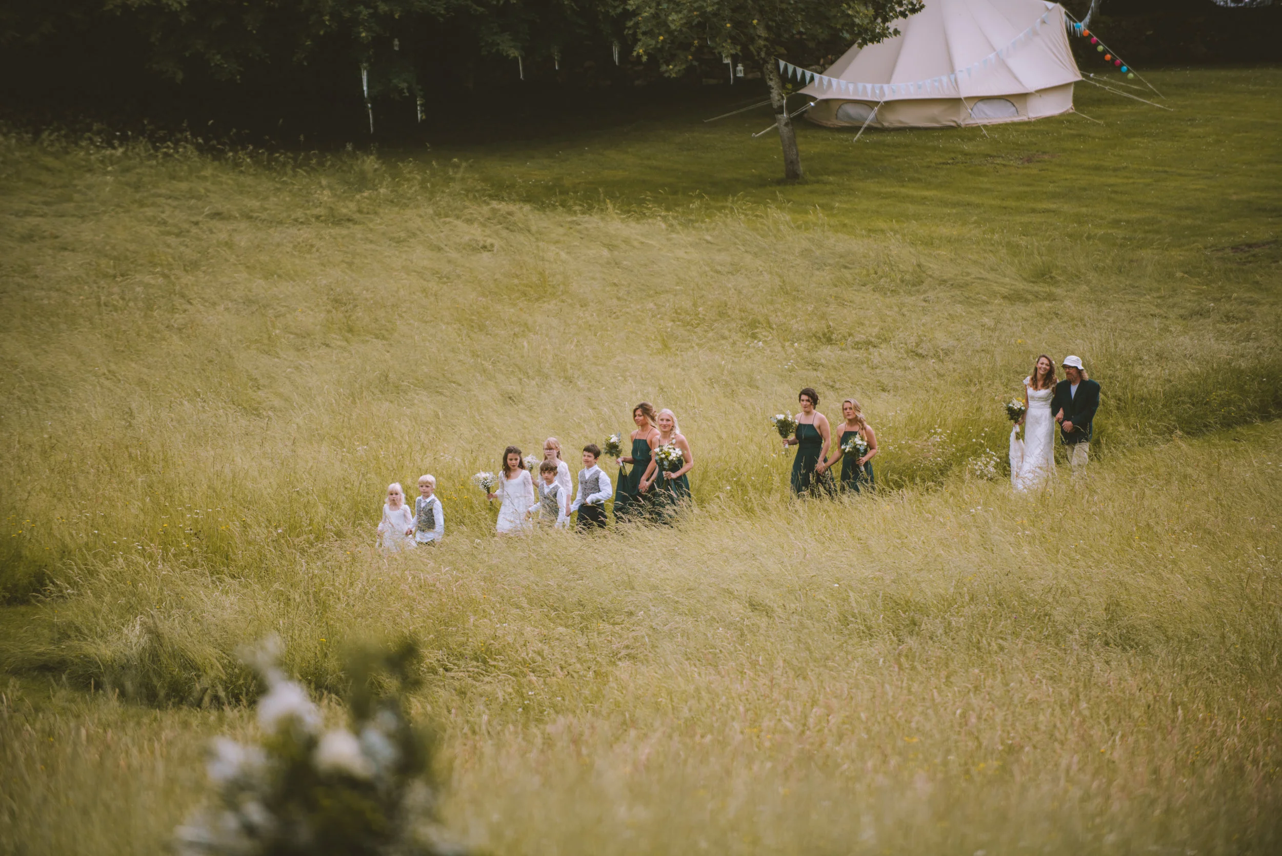 A wedding procession walking across a grassy field, with bridesmaids and flower girls in white and dark dresses, carrying flowers, observed by a couple in the distance, with a large tent in the background.
