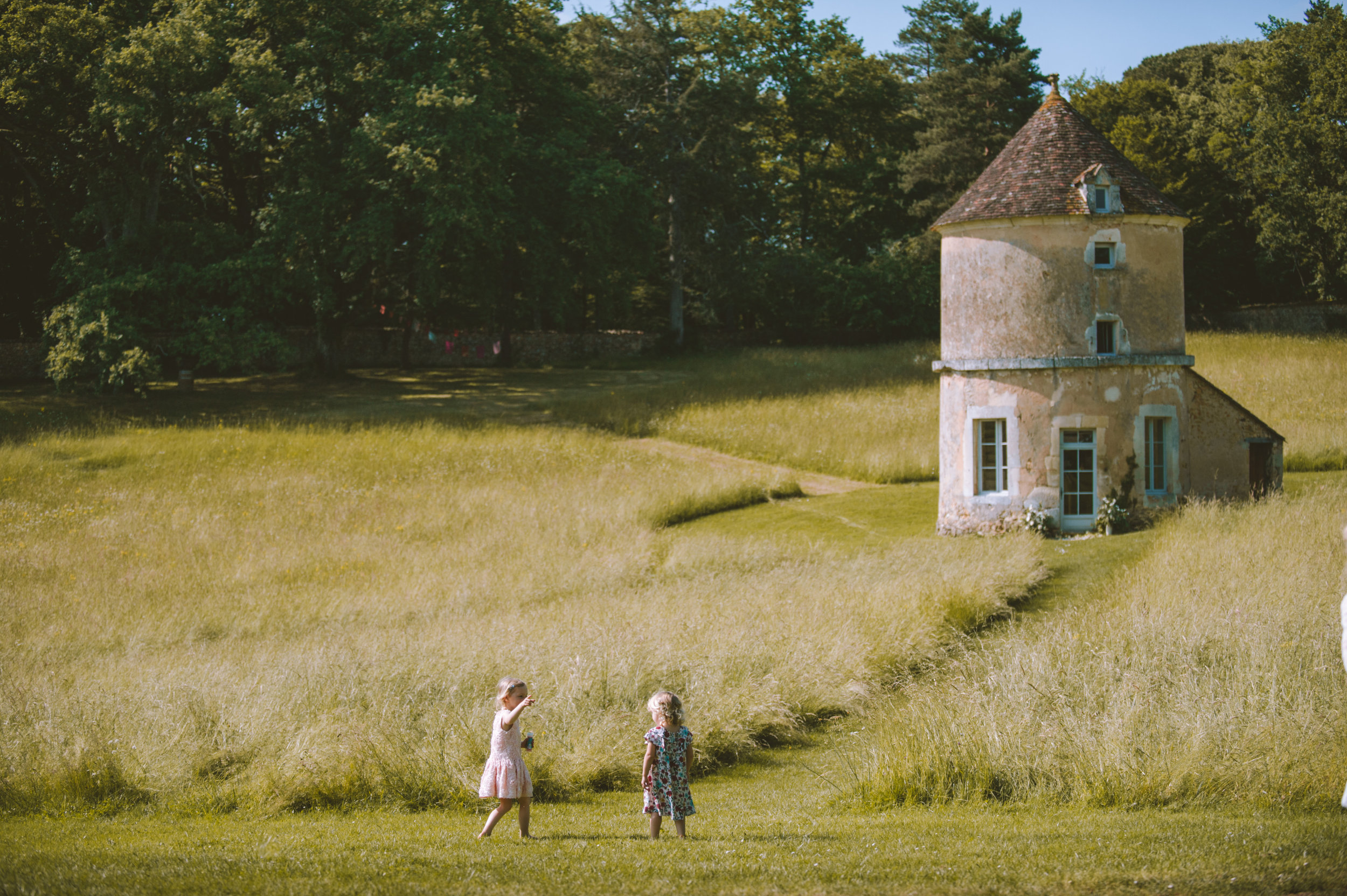 Two young girls standing in a grassy field, one pointing, with a small path leading to an old, weathered tower-like house surrounded by tall grass and trees.