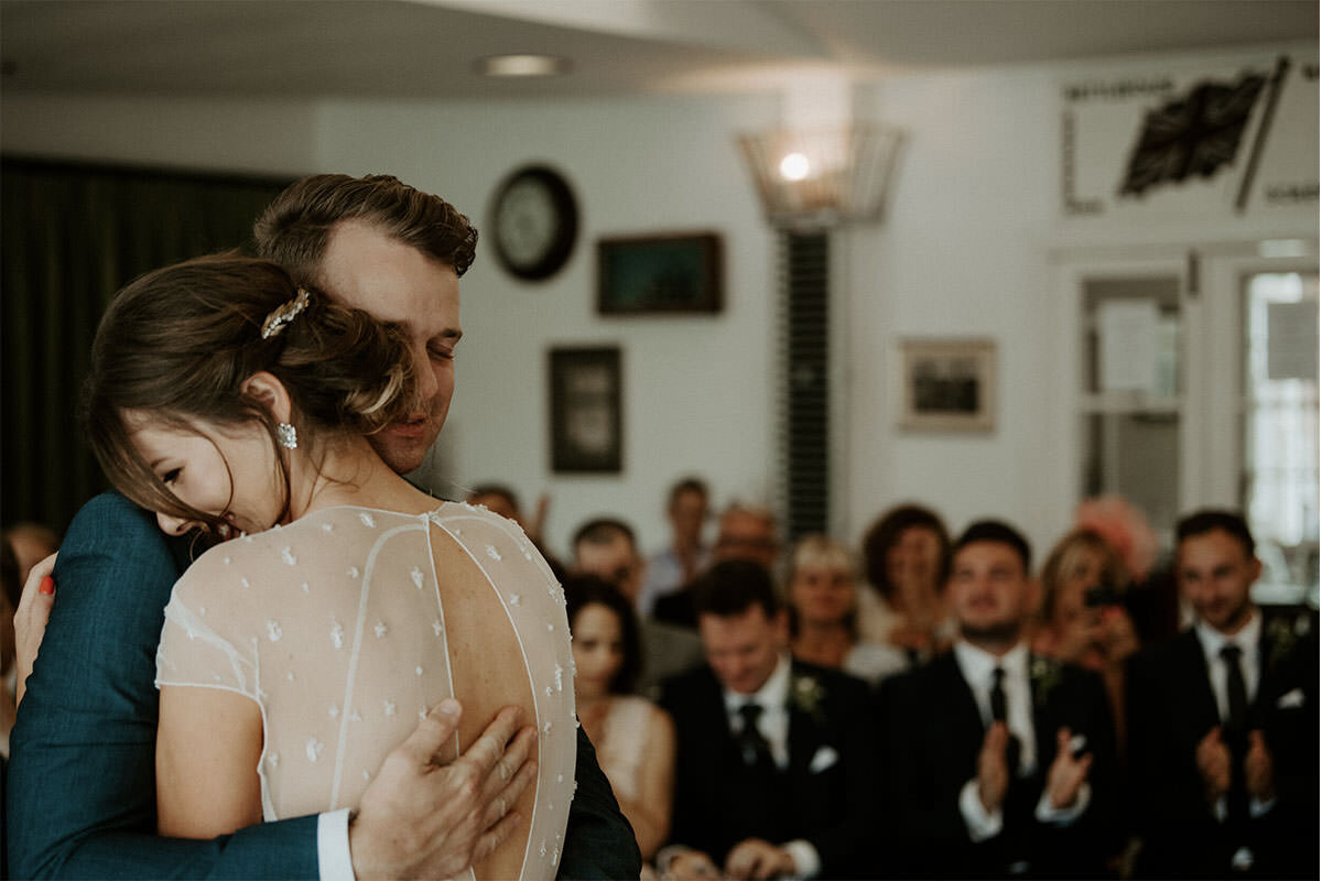 A bride and groom hugging during their wedding ceremony, with guests seated and clapping in the background.