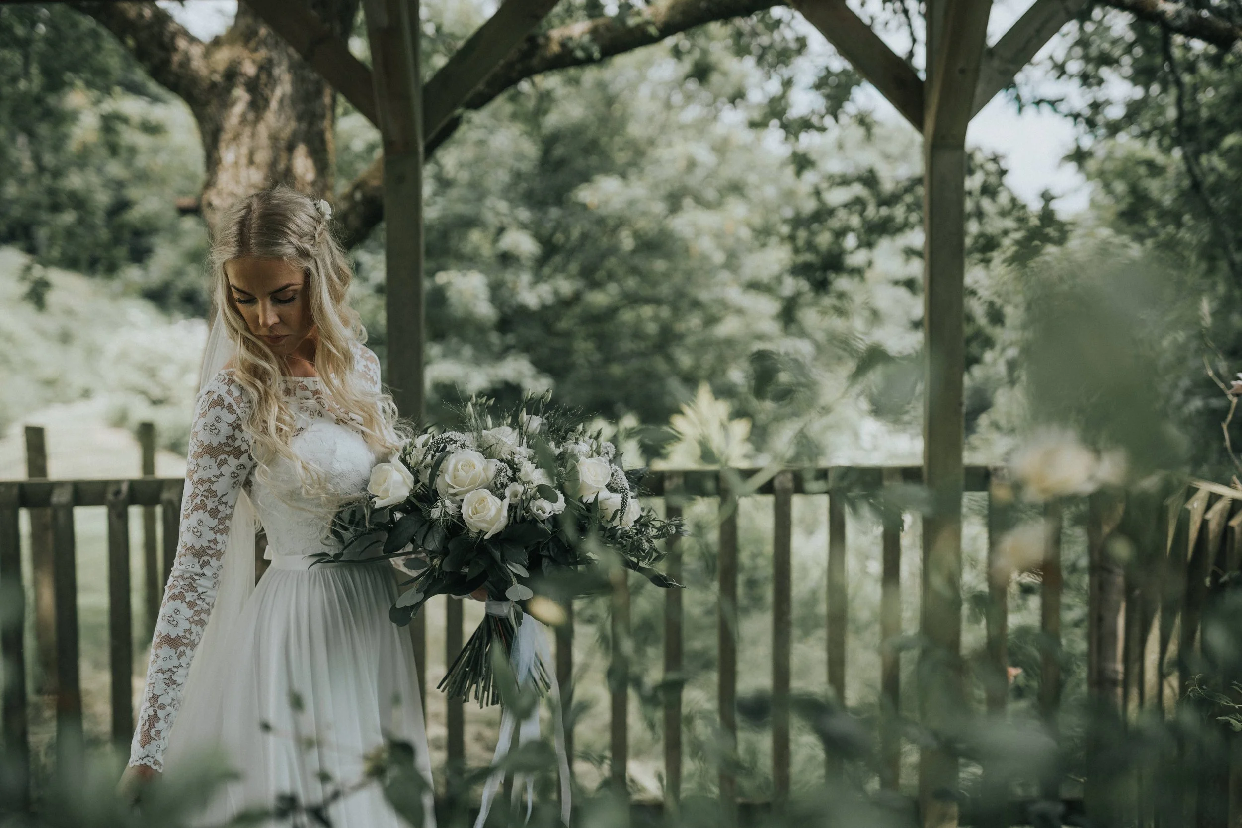 A bride in a white lace wedding dress holding a bouquet of white roses and greenery stands outdoors under a wooden gazebo, surrounded by trees and greenery.