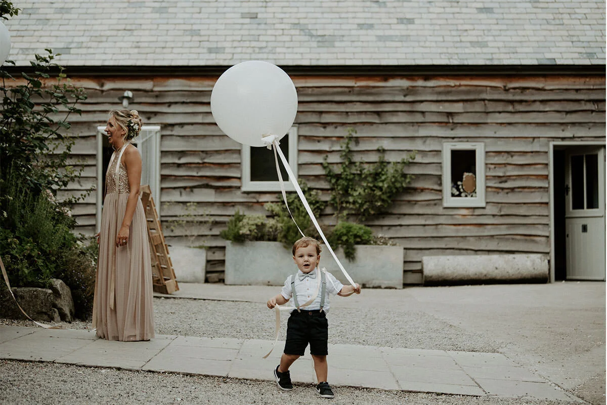 A young boy holding a large white balloon outside a rustic wooden building with a woman in a beige dress smiling nearby.
