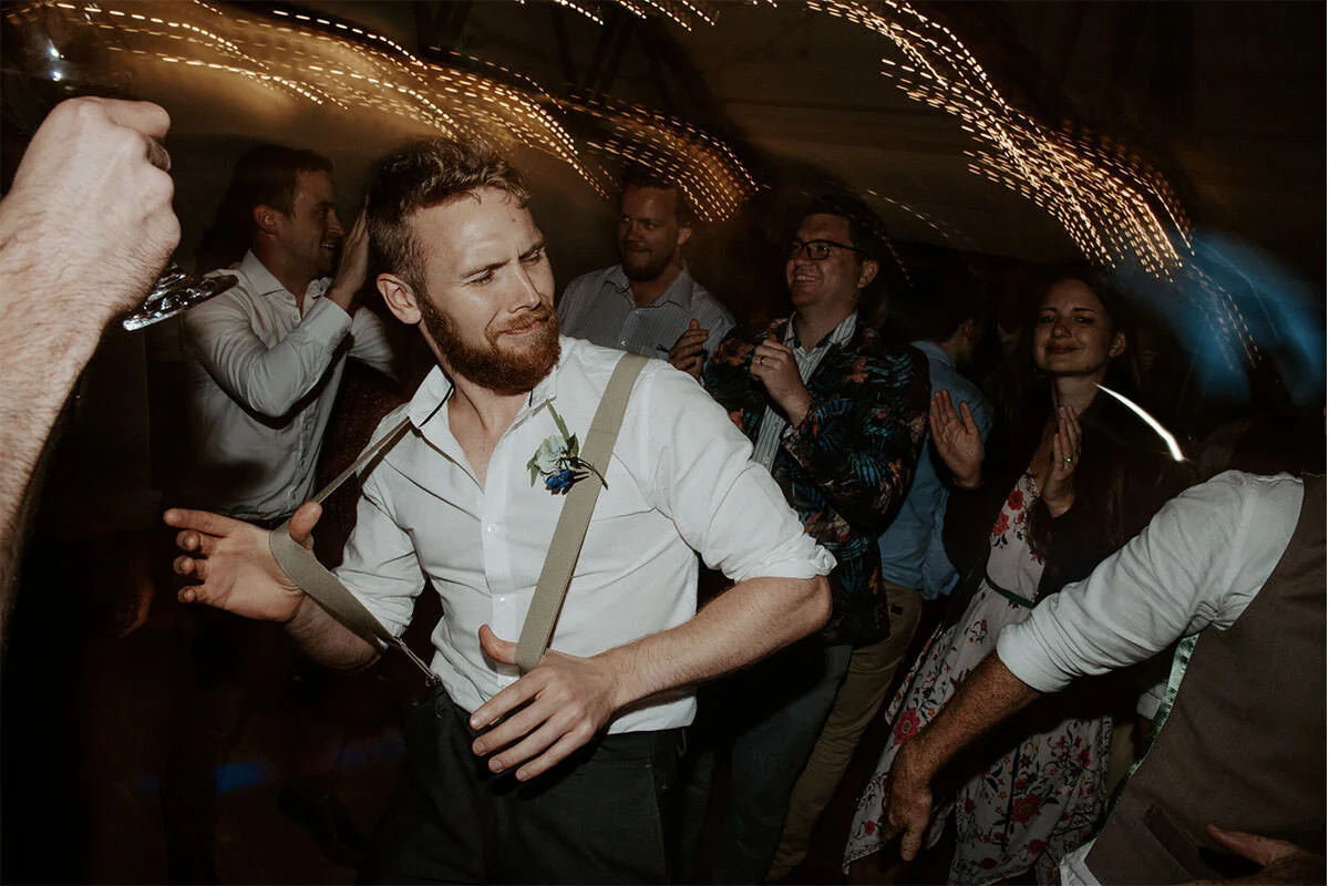 Man with suspenders and a boutonniere dancing at a party with other guests clapping and enjoying the music.