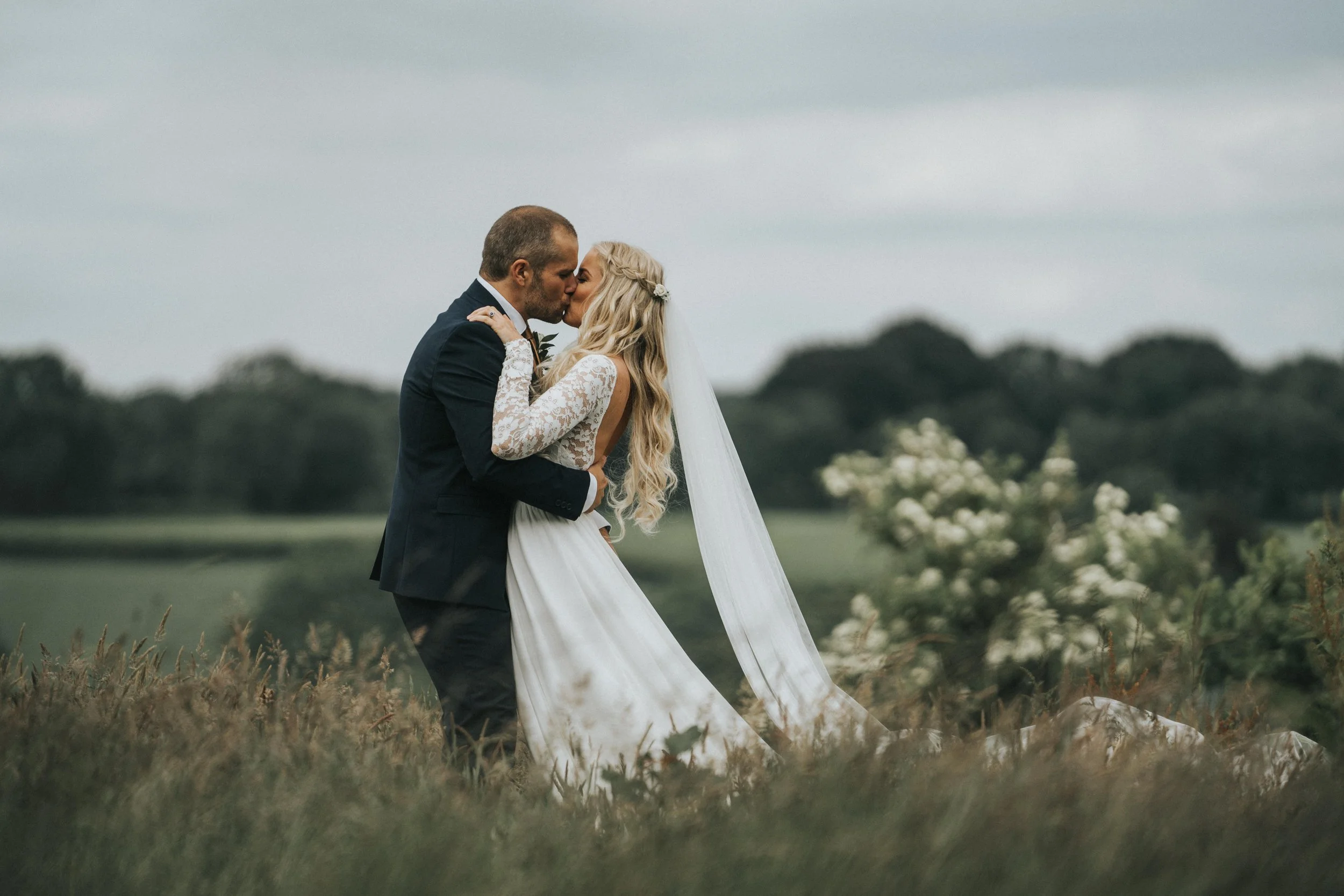 A newlywed couple sharing a romantic kiss outdoors in a field, with the bride in a white lace wedding gown and long veil, and the groom in a dark suit, against a backdrop of trees and cloudy sky.