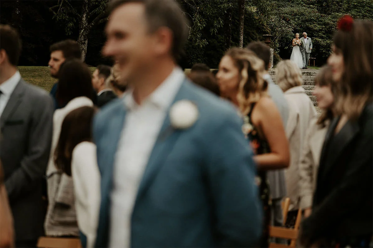 Wedding scene with guests standing outdoors, a bride and groom walking up steps in the background.