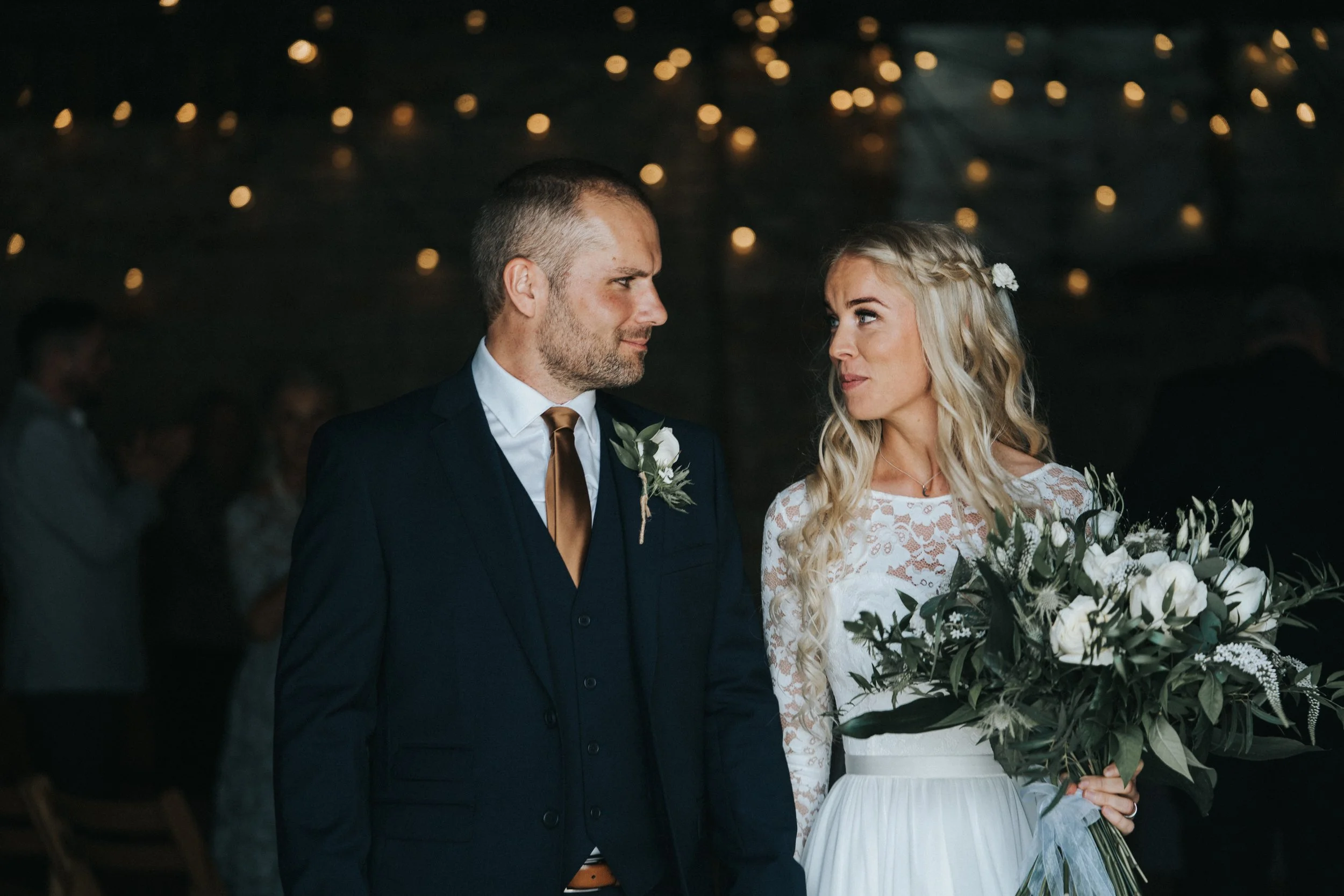 A bride and groom standing inside a dimly lit wedding venue with string lights overhead, looking at each other. The bride holds a large bouquet of white flowers and greenery. The groom wears a dark suit with a white shirt and brown tie.