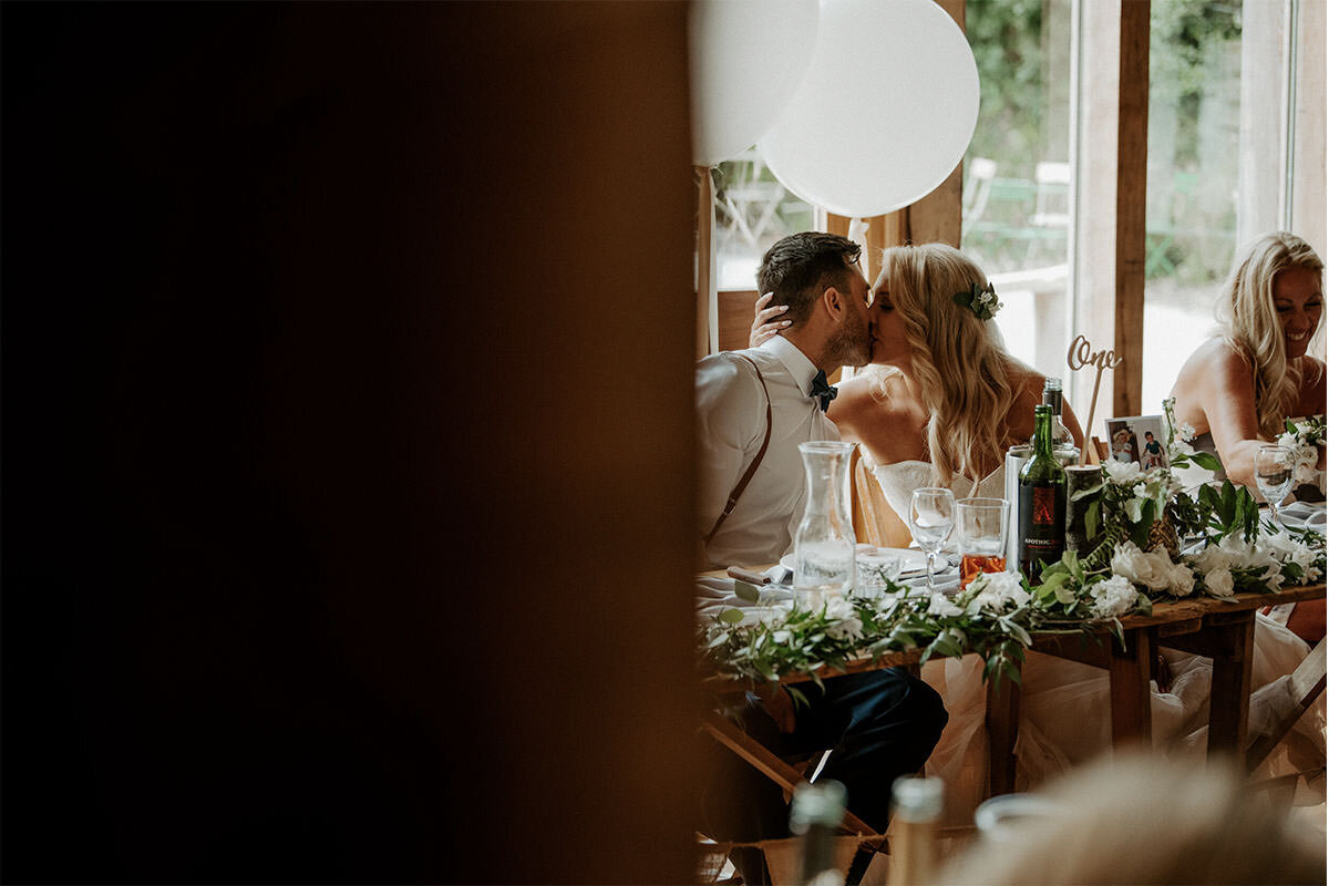 A bride and groom kiss during a wedding reception, seated at a decorated table with drinks and floral arrangements.