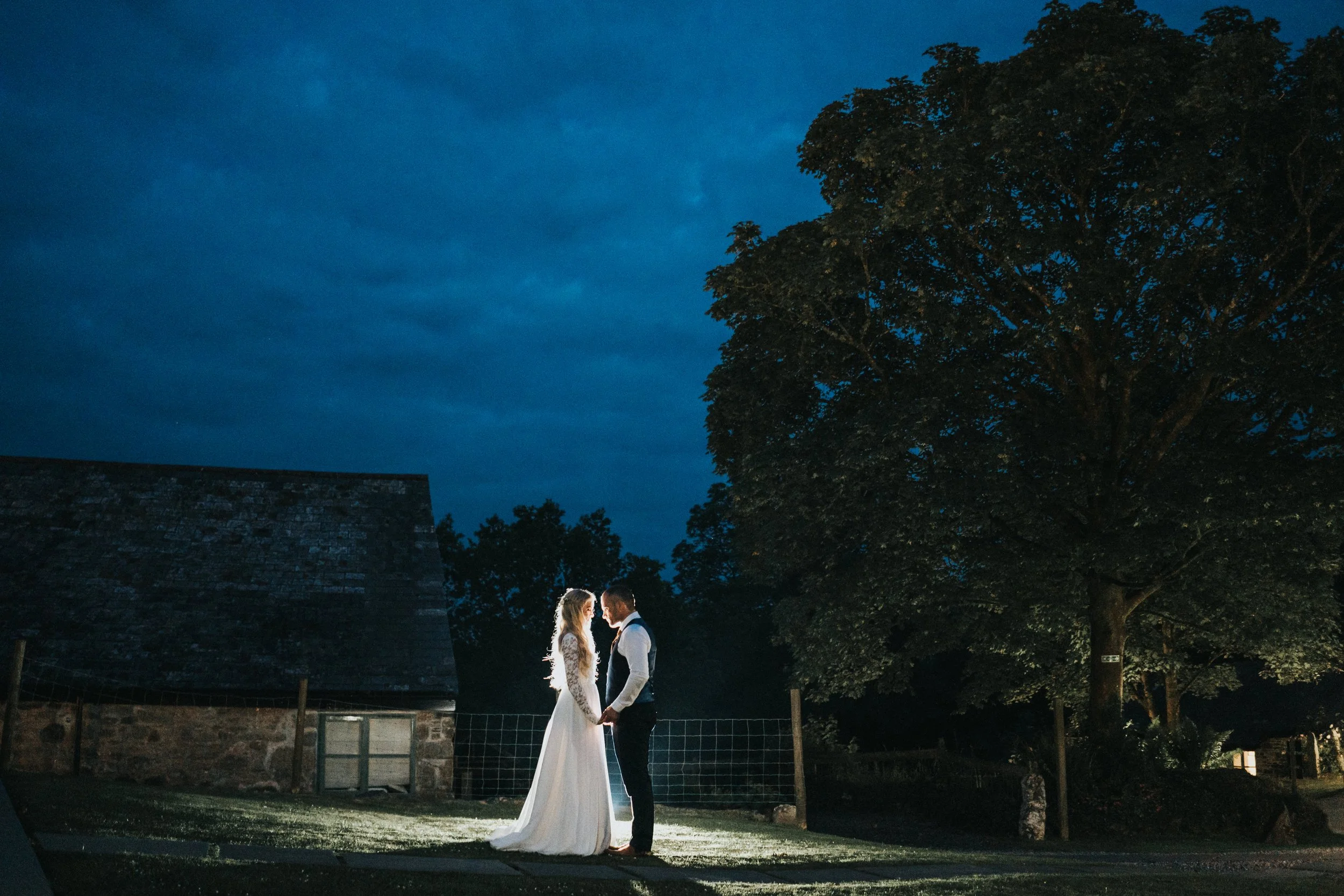 A bride and groom standing close, holding hands and facing each other outdoors at night, illuminated by a spotlight, with a large tree and a rustic building in the background.