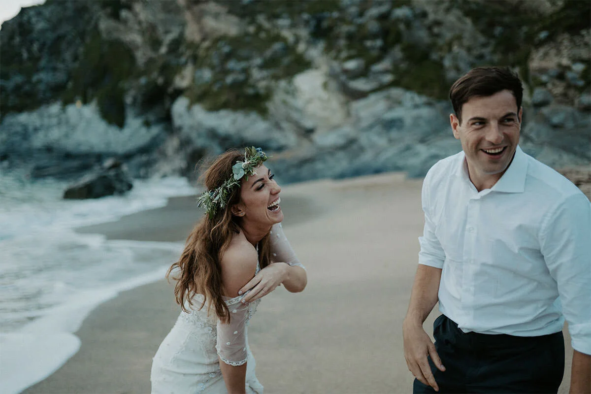 A man and woman laughing and enjoying themselves on a beach with rocks and water in the background. The woman is wearing a floral crown and white dress, and the man is dressed in a white shirt and dark pants.
