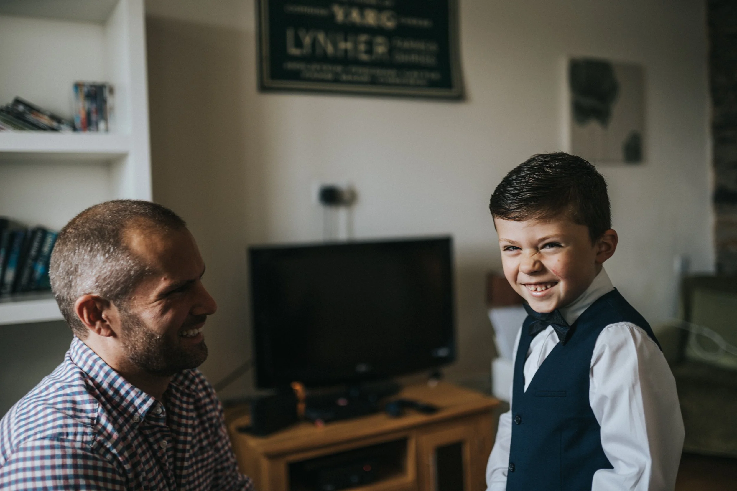 A man and a boy sharing a joyful moment indoors, with the boy making a funny face and the man smiling, in a living room with a television and books.