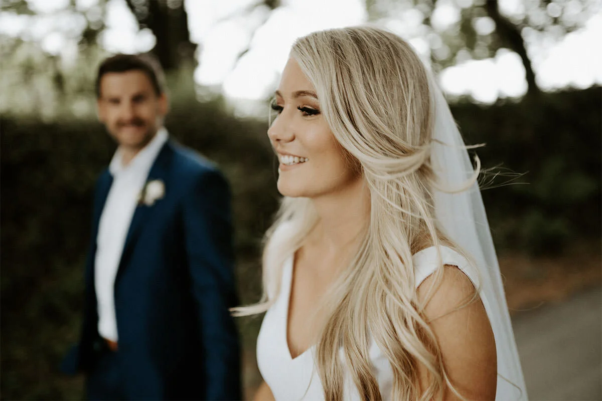A bride smiling with her eyes closed standing outdoors, with a groom blurred in the background, trees and natural scenery behind them.