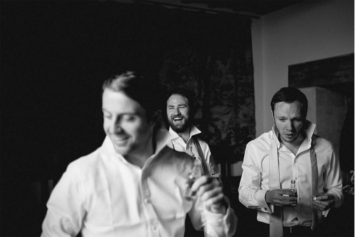Three men dressed in white shirts with suspenders, socializing and drinking alcohol in a dimly lit setting, likely a bar or a party, with one person smiling broadly.