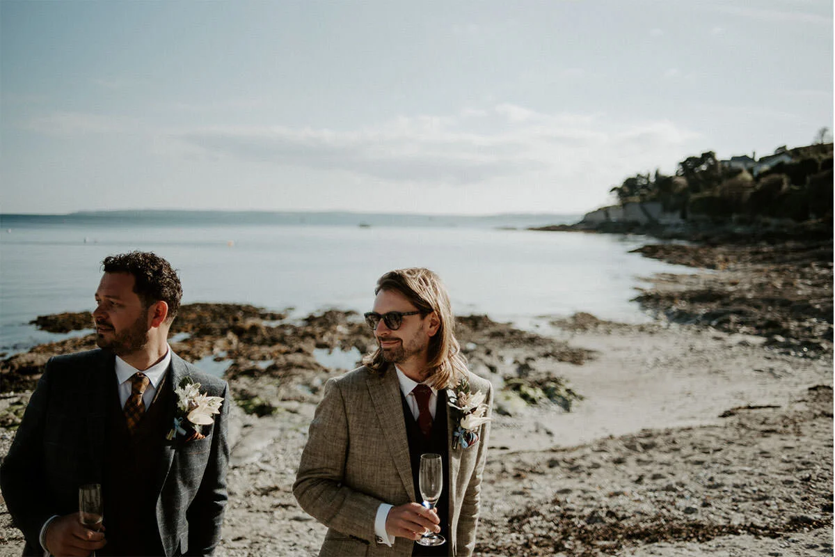 Two men in suits with boutonnières and glasses of champagne standing on a rocky beach, with water and a distant shoreline in the background.