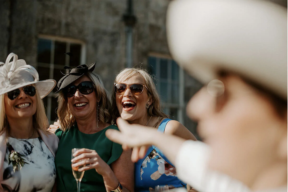 Three women wearing large hats and sunglasses smiling and laughing together at an outdoor event, with a fourth person partially visible in the foreground.