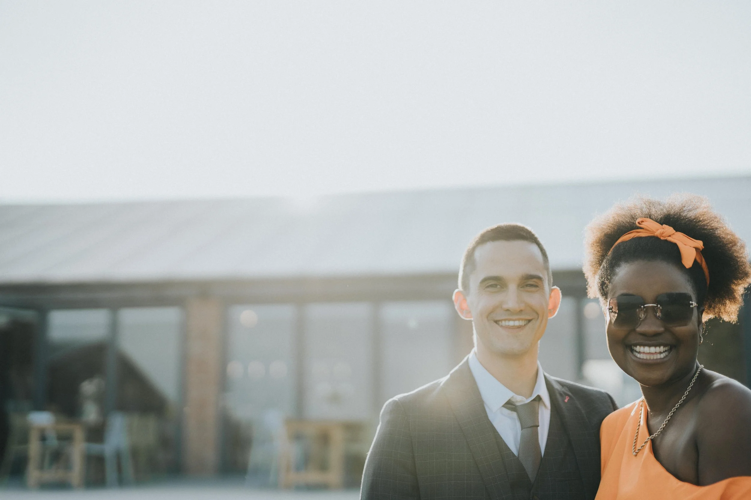 A smiling man in a suit and a woman wearing sunglasses and an orange dress standing outdoors.