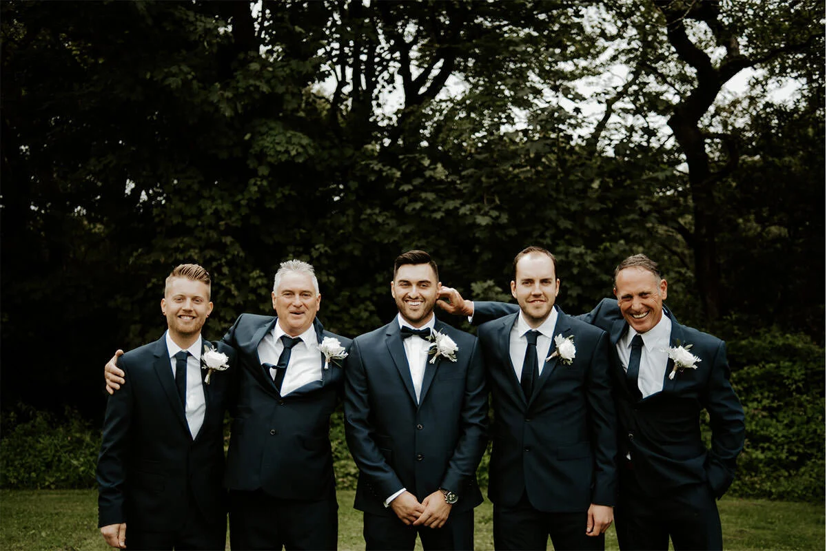 Group of five men in suits and ties, standing outdoors with trees in background, smiling for a photo at a formal event.