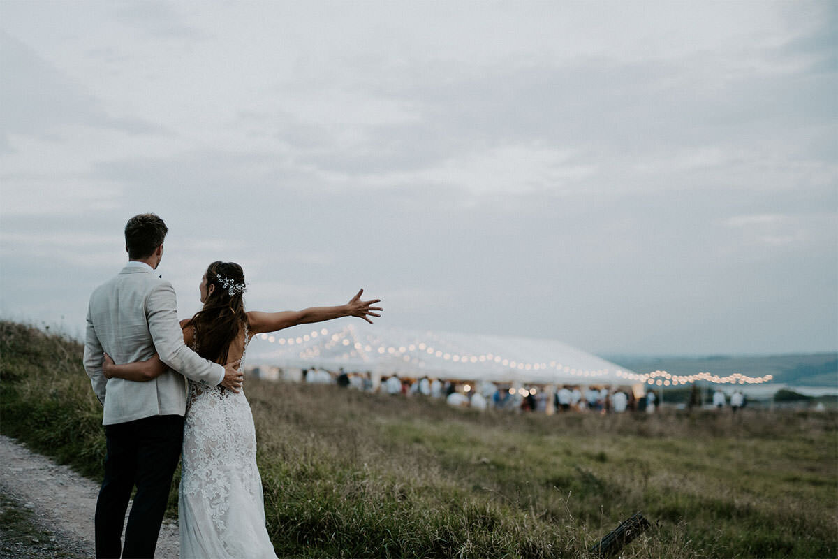 A bride and groom standing outdoors near the beach during a wedding, with the bride extending her arm as if waving or reaching out, and wedding guests in the background under string lights.