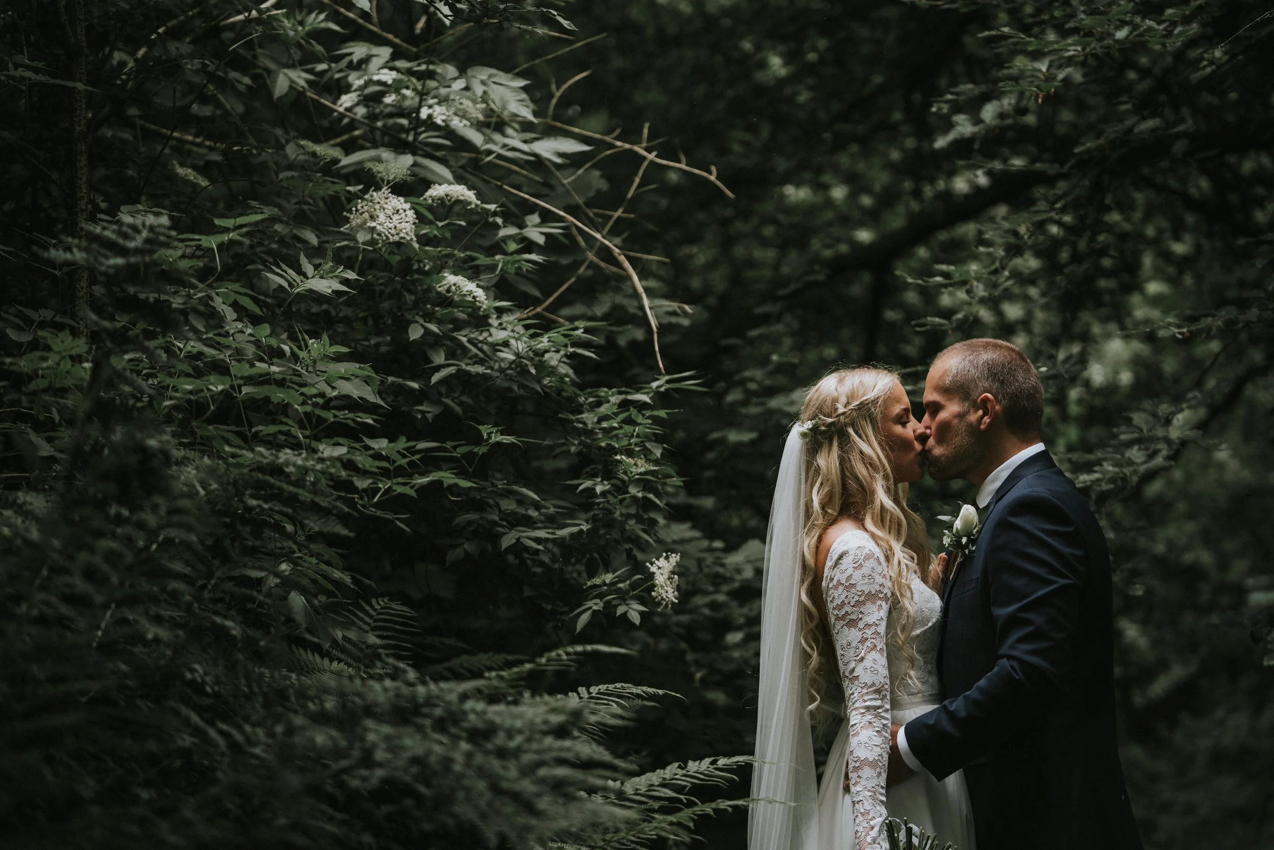 A bride and groom share a kiss in a wooded setting, surrounded by lush green foliage.