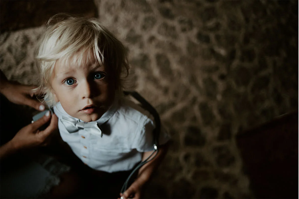 A young blonde boy with blue eyes looking up at the camera, wearing a white shirt, with someone adjusting his bow tie.