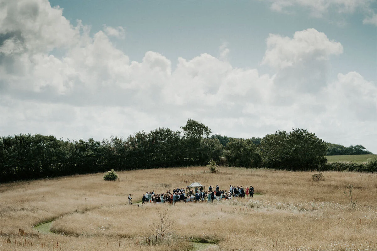 A group of people gathered in an open grassy field with trees in the background under a partly cloudy sky.