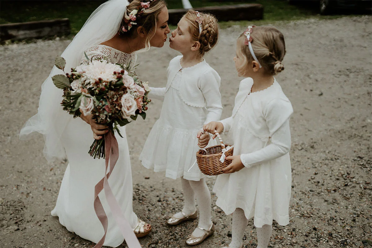 A bride leaning down to kiss two young girls at an outdoor wedding. The bride holds a large bouquet, and one girl holds a small wicker basket. All are dressed in white, with floral headbands, on a gravel path.
