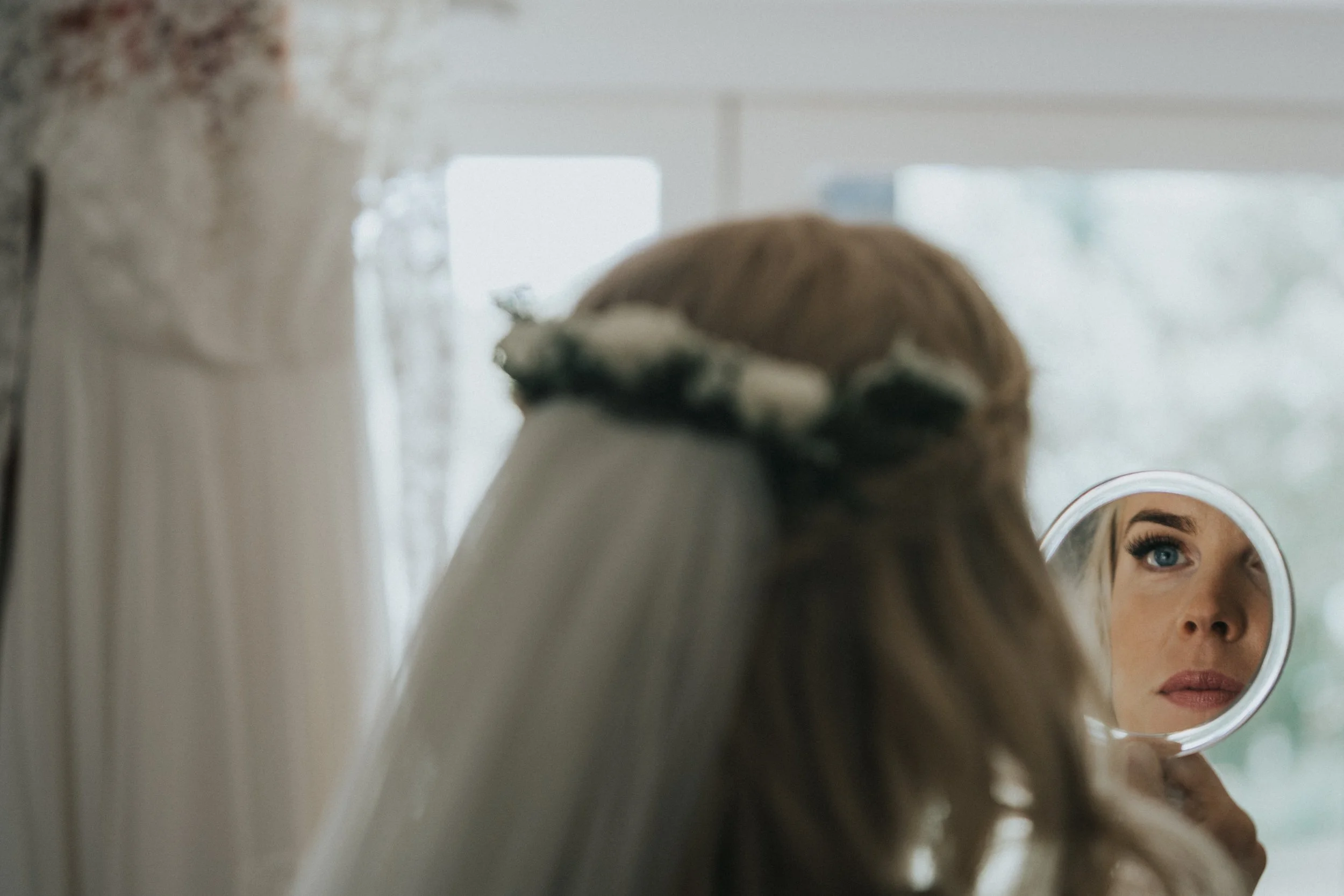 A woman with blonde hair wearing a flower crown looks into a handheld mirror, reflecting her face with blue eyes and makeup, while standing in a room with natural light.