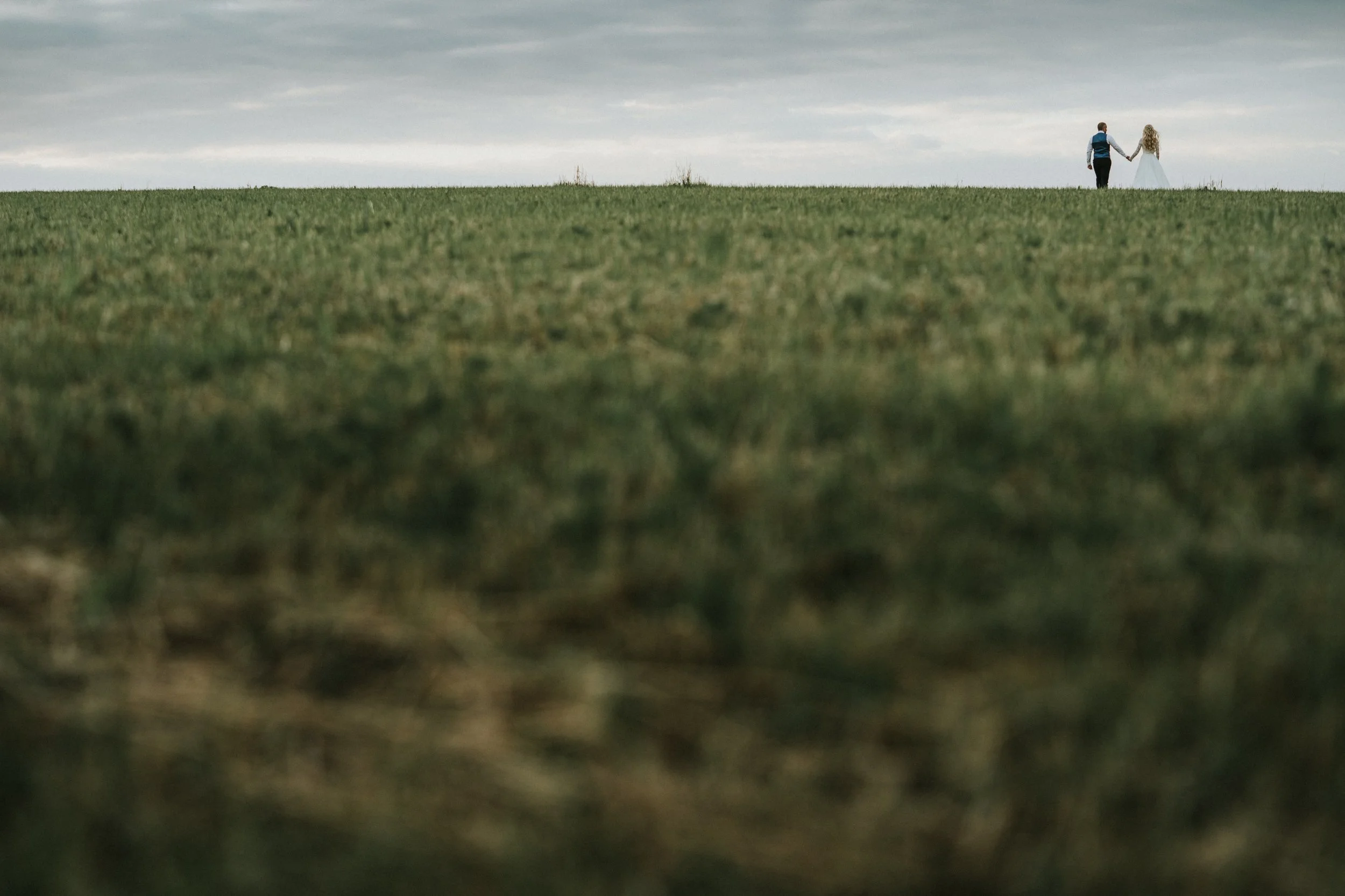 A couple walking hand in hand in a grassy field on a cloudy day.